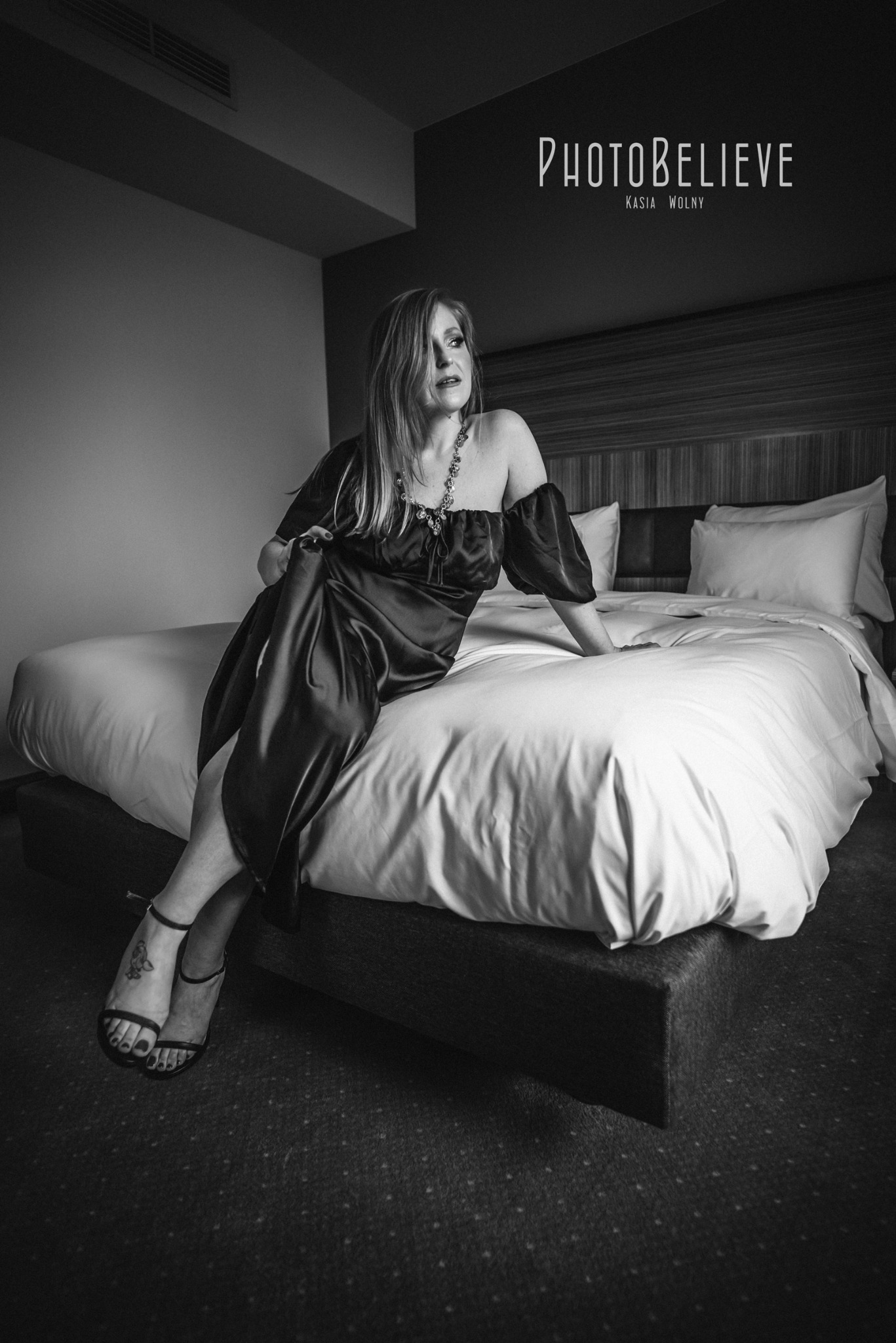 Czarno-białe zdjęcie kobiety w satynowej sukni i sandałach na obcasie, siedzącej na łóżku w pokoju hotelowym, z widocznym logo fotografa w górnym rogu.