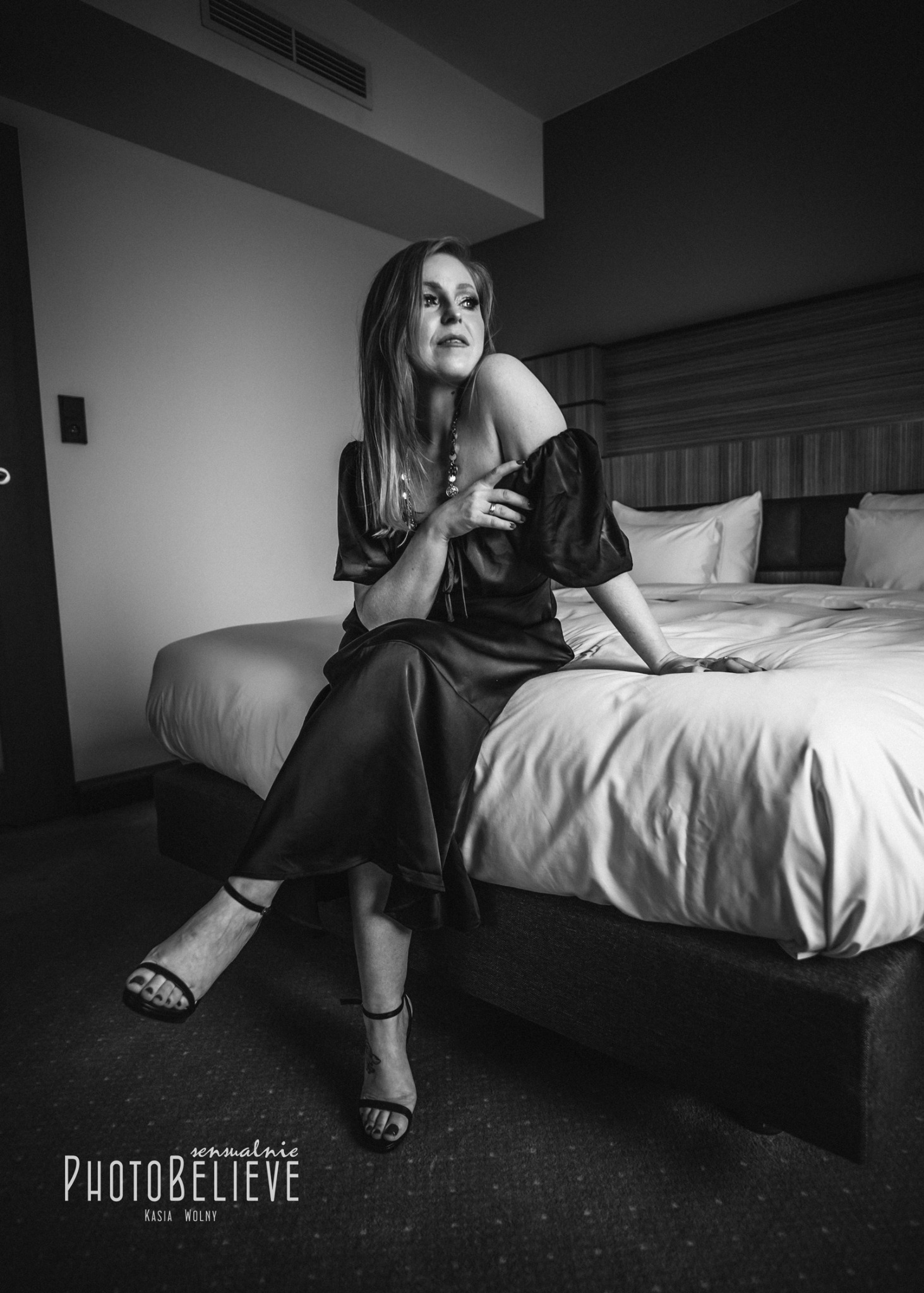 Czarno-białe zdjęcie kobiety w satynowej sukience siedzącej na łóżku w pokoju hotelowym, z widocznym logo fotografa w rogu.
