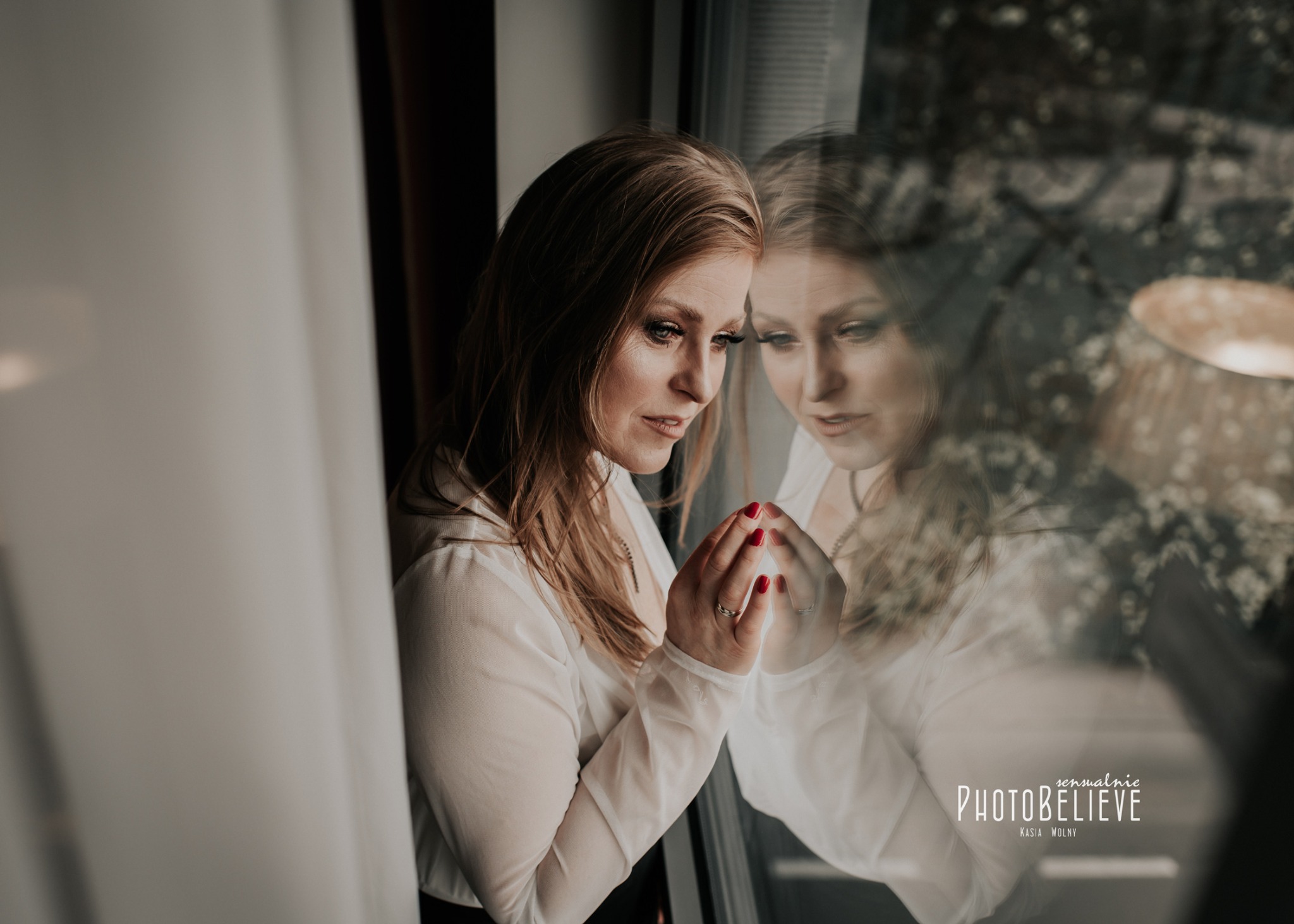 Kobieta w białej koszuli opiera dłonie o szybę okna, widoczne odbicie postaci i fragment lampy w tle.