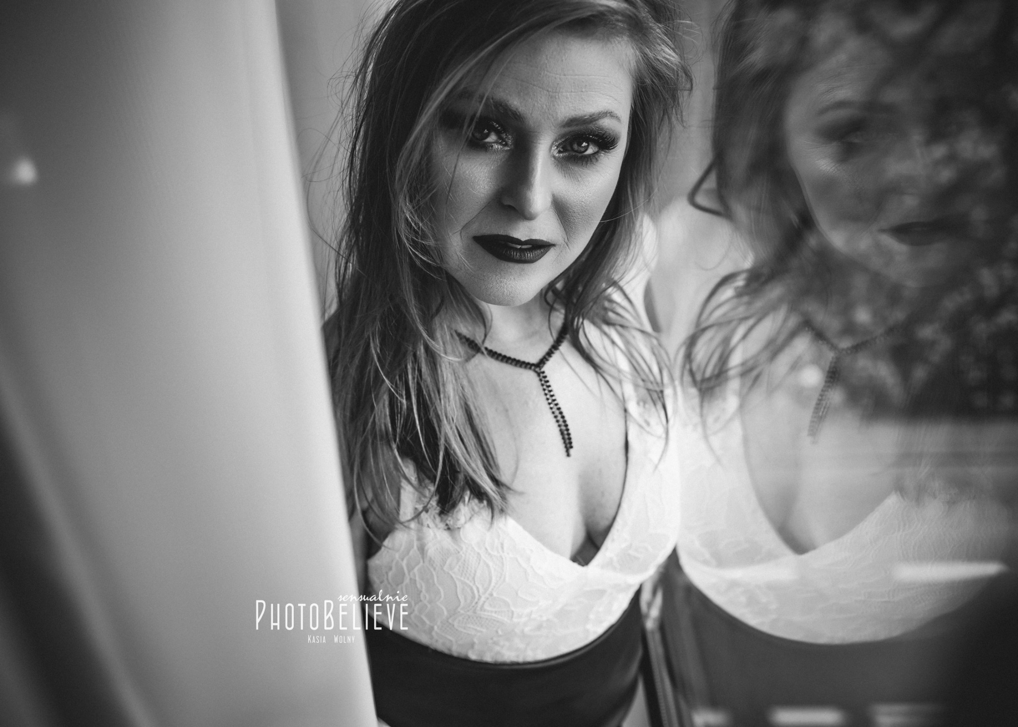 Portret kobiety z mocnym makijażem oczu i ust, ubrana w koronkową bluzkę z głębokim dekoltem, widoczny napis 'PhotoBelieve' w lewym dolnym rogu, odbicie twarzy w tle.