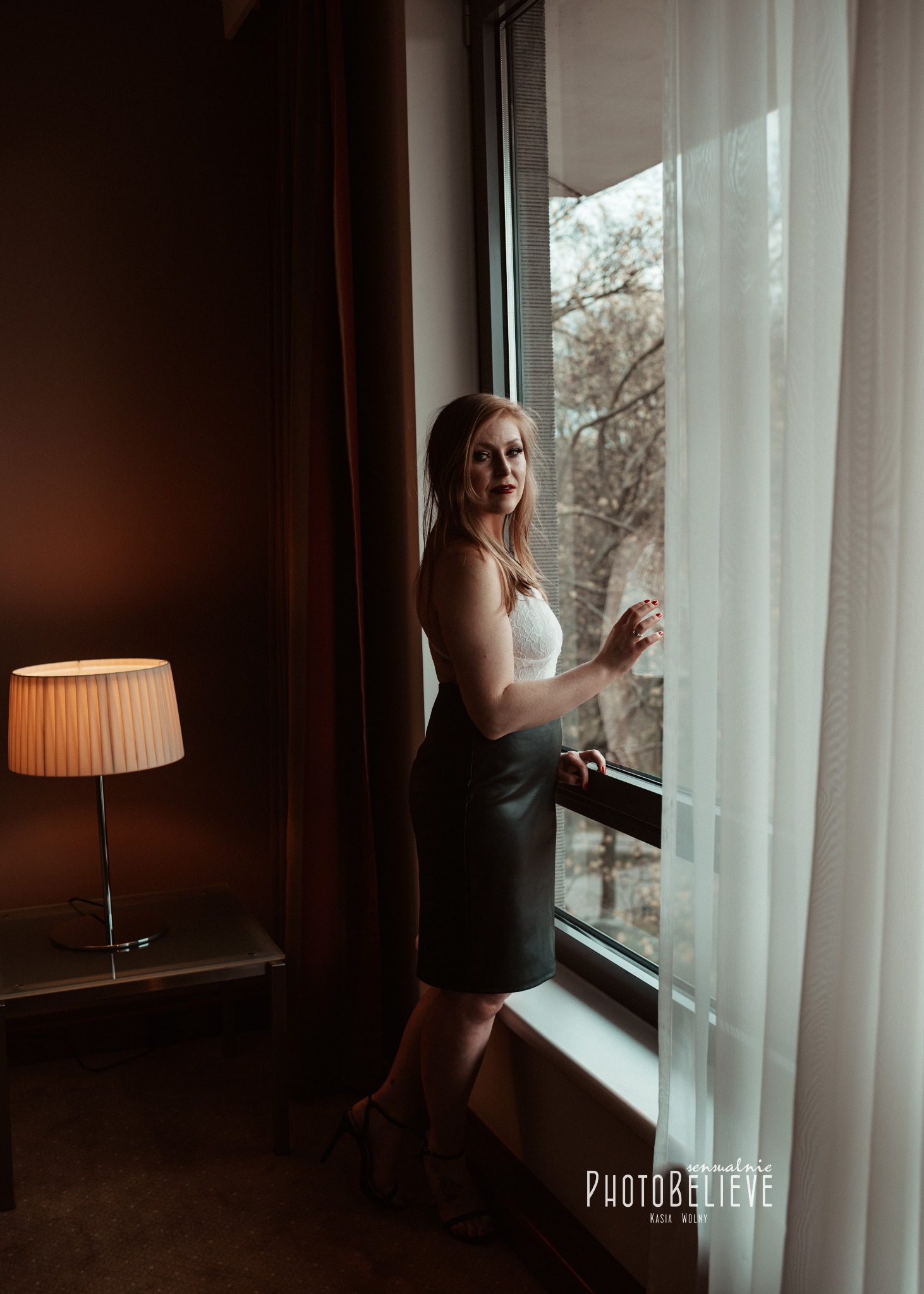 Kobieta w koronkowej bieliźnie i skórzanej spódnicy stoi przy oknie, spoglądając na zewnątrz, w pokoju z lampą na stoliku nocnym.