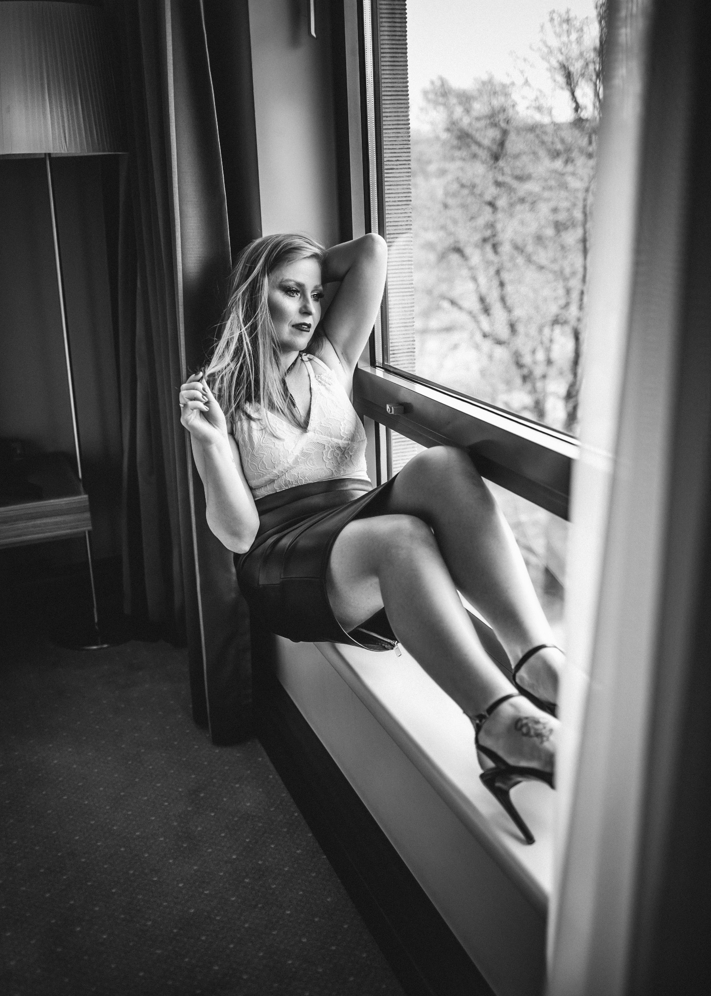 Czarno-białe zdjęcie kobiety siedzącej na parapecie okna, ubrana w koronkową bluzkę i skórzaną spódnicę, z rękami uniesionymi za głową, patrzącej na zewnątrz.