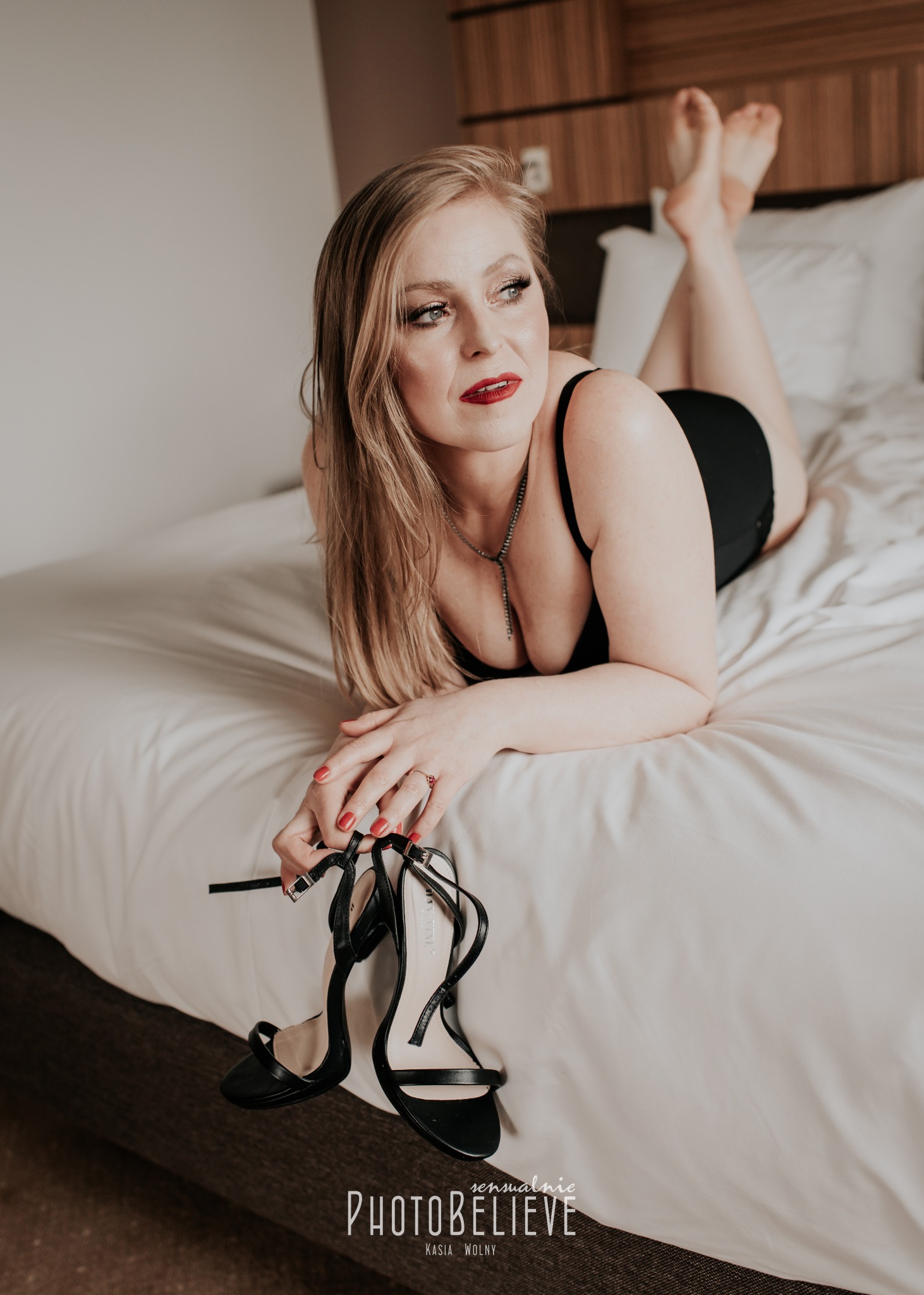 Kobieta w czarnej bieliźnie leży na łóżku, trzymając czarne szpilki, z pomalowanymi na czerwono paznokciami i delikatną biżuterią.