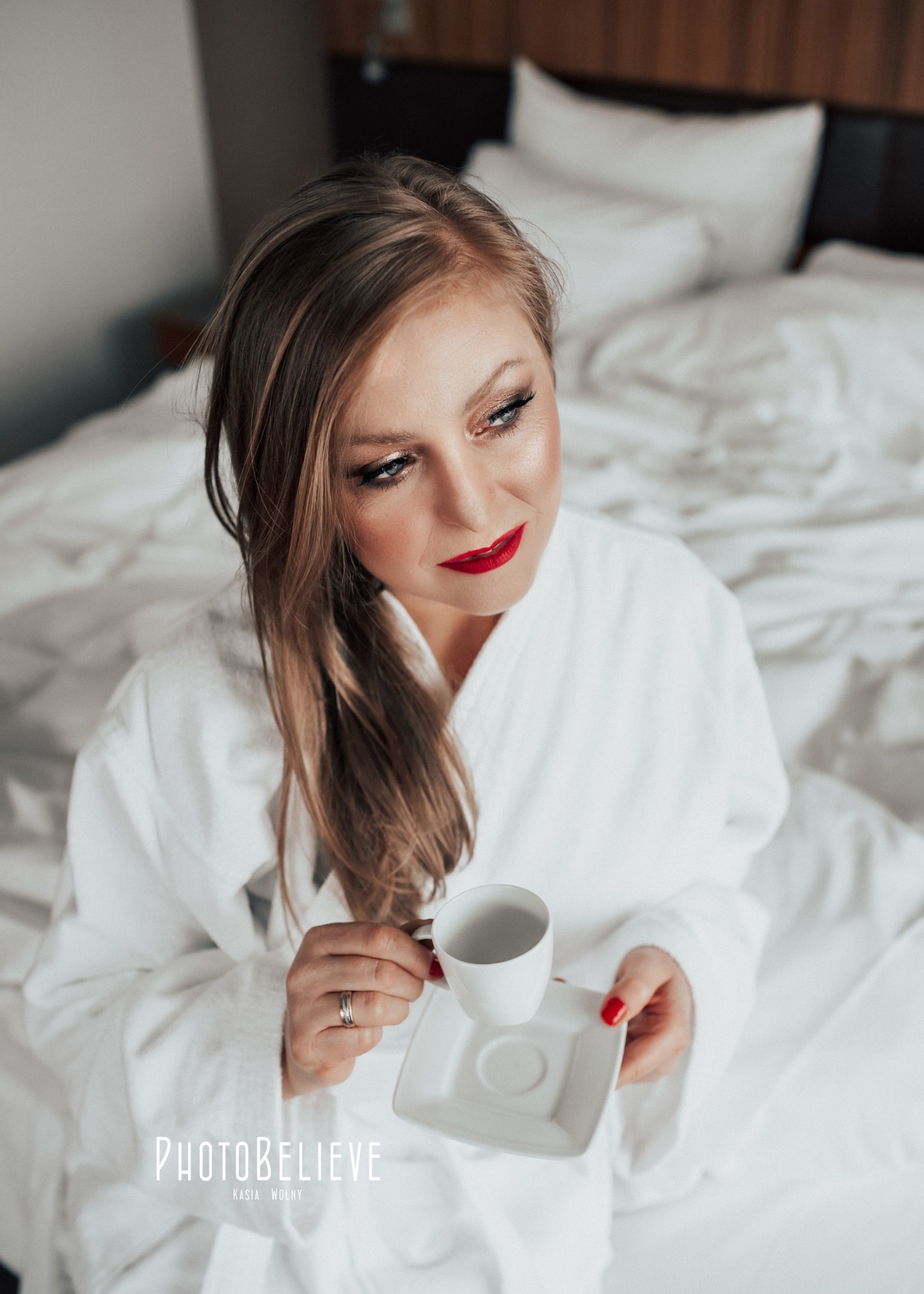 Kobieta w białym szlafroku siedzi na łóżku z filiżanką kawy, makijaż z czerwonymi ustami, spojrzenie skierowane w górę.
