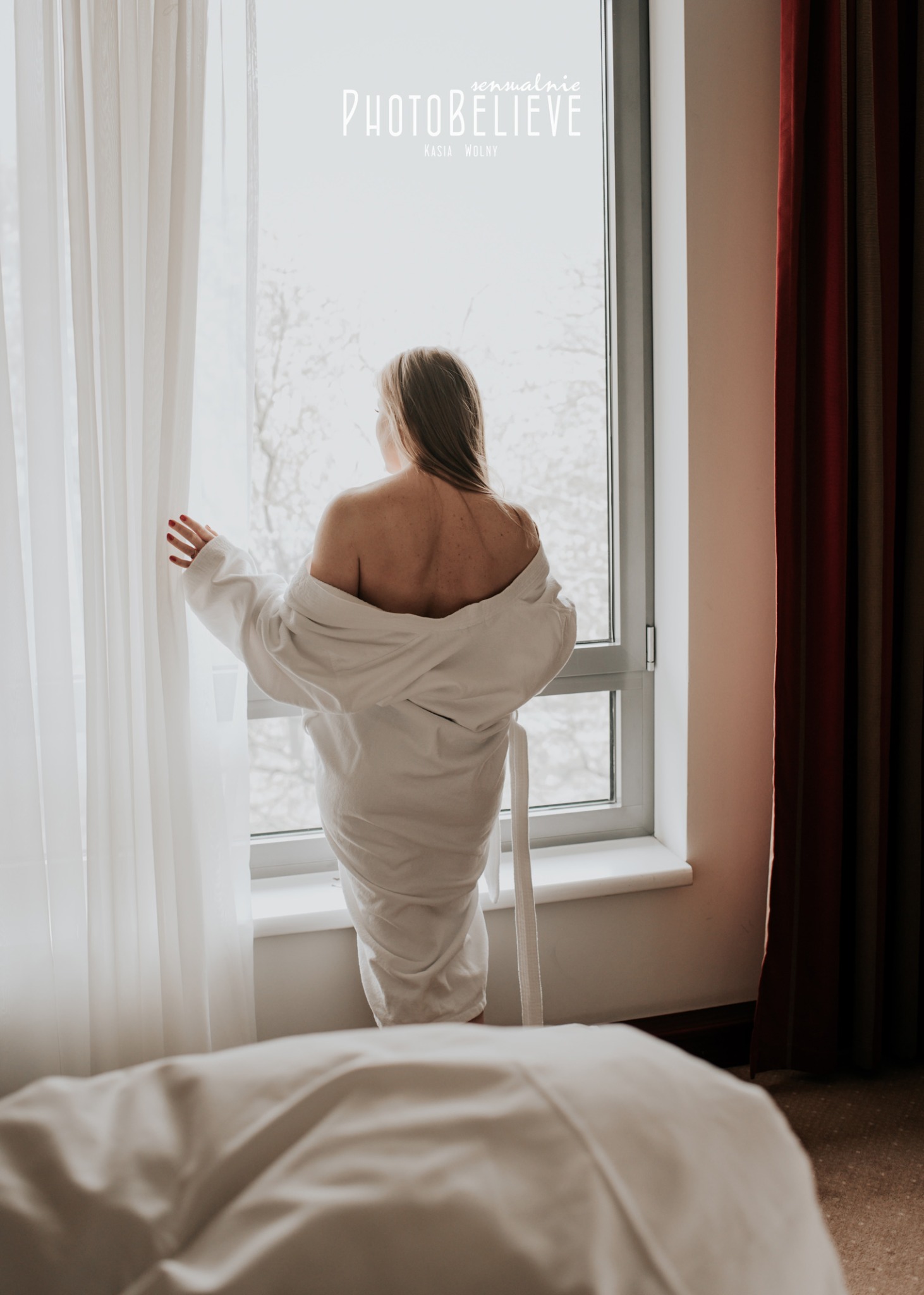 Kobieta w białym szlafroku stoi tyłem przy oknie, spoglądając na zewnątrz, z widocznym logo fotografa w górnej części kadru.