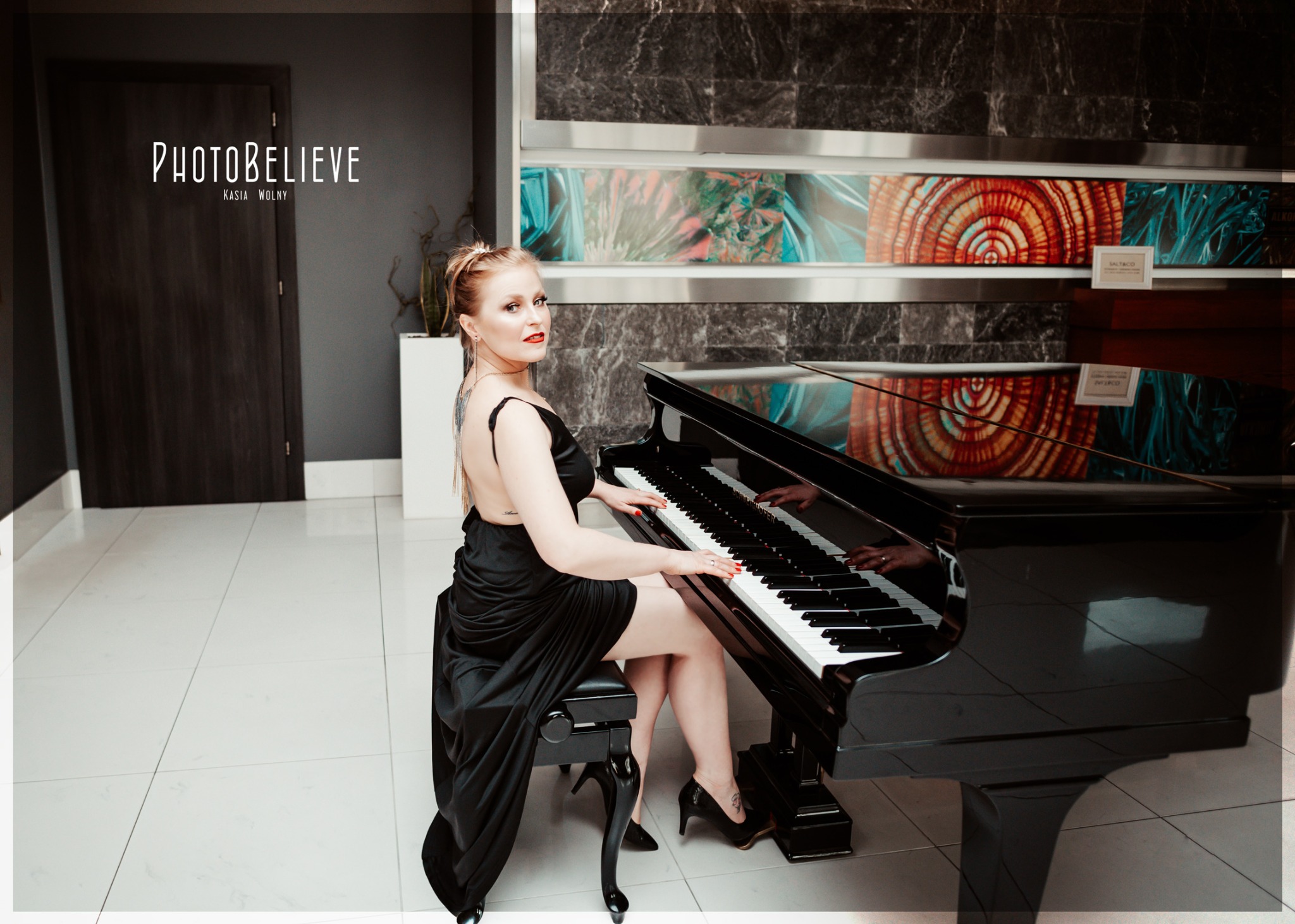 Elegancka kobieta w czarnej sukni gra na czarnym fortepianie w nowoczesnym wnętrzu z marmurową ścianą i dekoracyjnymi panelami.