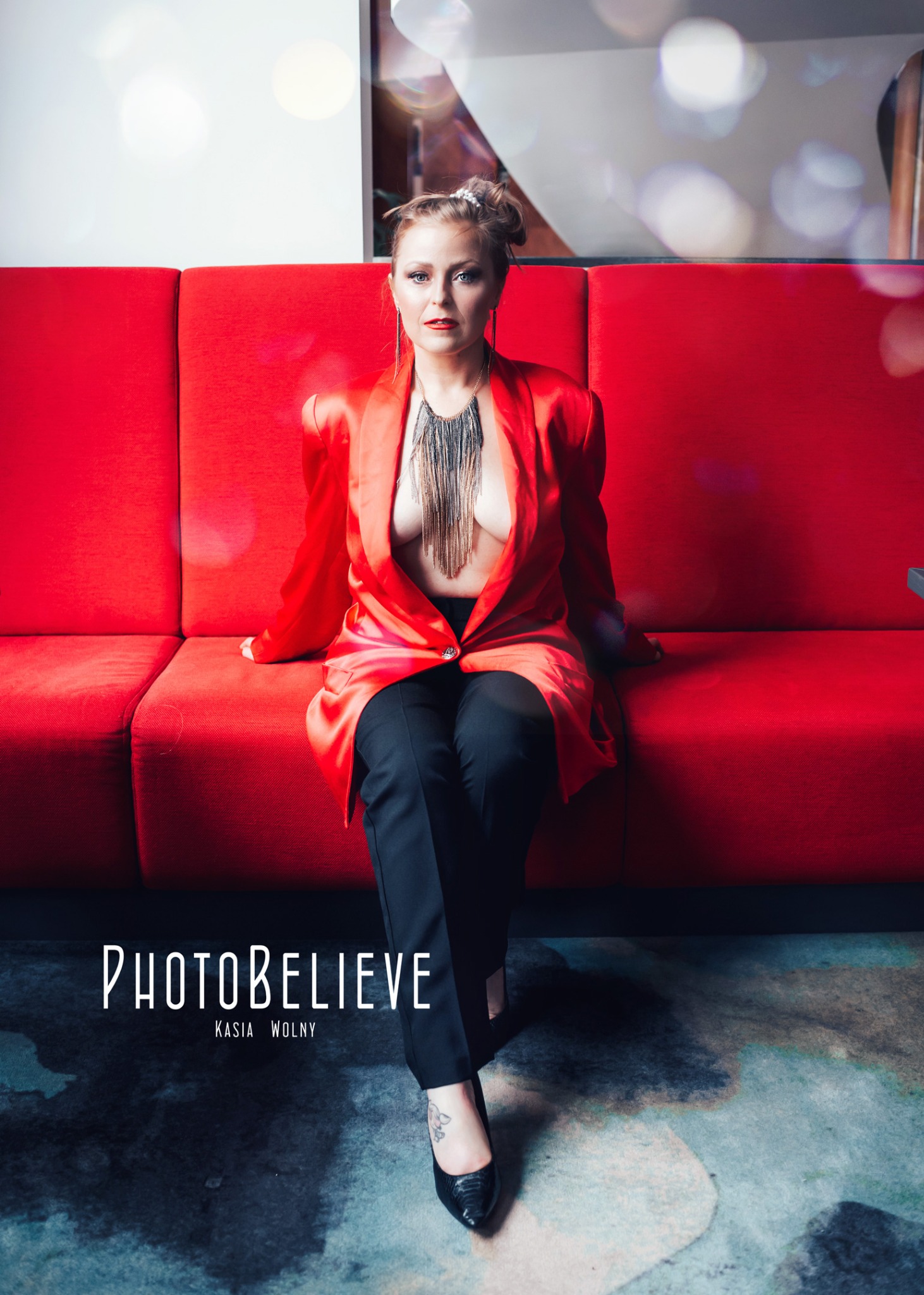 Portret kobiety siedzącej na czerwonej sofie, ubrana w satynową marynarkę i czarne spodnie, z ozdobnym naszyjnikiem.