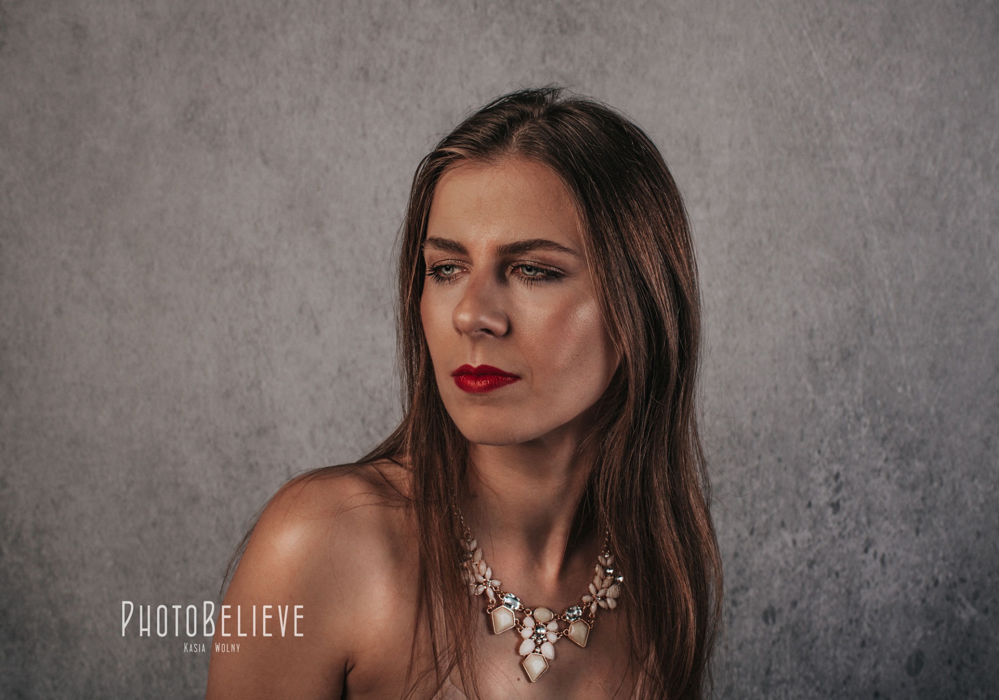 Portret kobiety z długimi brązowymi włosami, w wyrazistym makijażu i ozdobnym naszyjniku na tle szarej ściany.