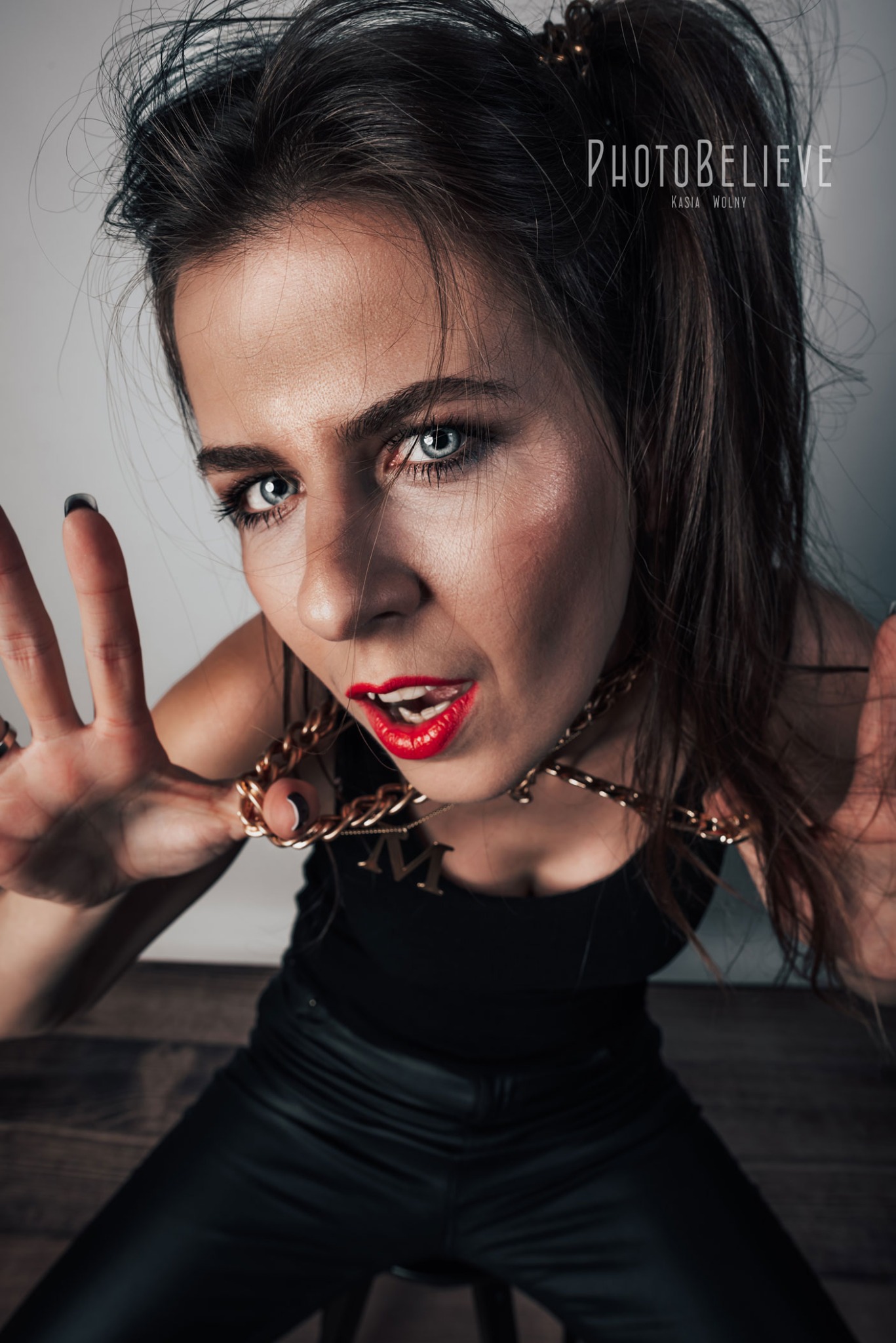 Portret kobiety z mocnym makijażem, w czarnym stroju i złotej biżuterii, uchwycony z perspektywy żabiej, z wyciągniętymi dłońmi w kierunku obiektywu.