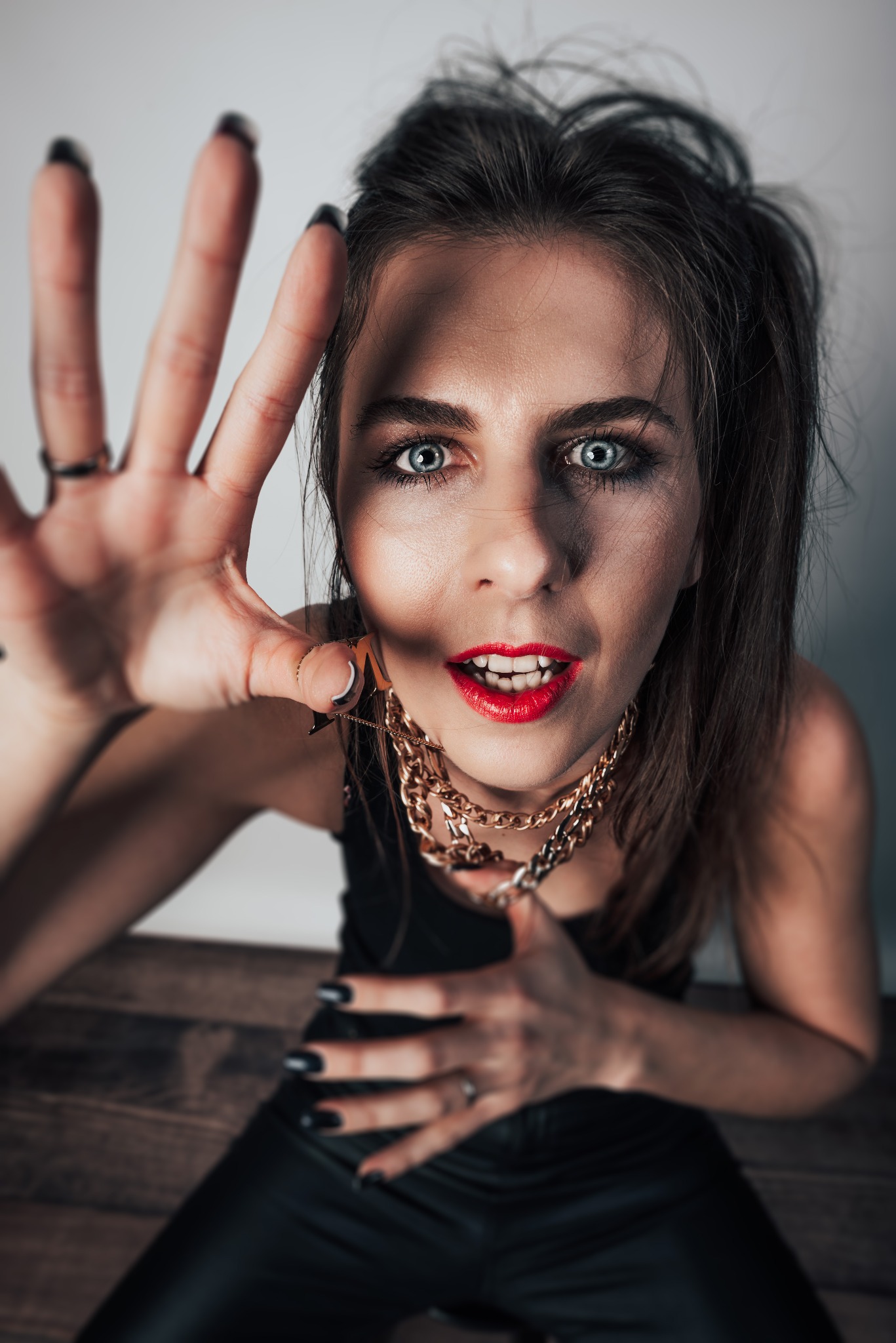 Portret kobiety z wyrazistym makijażem, złotym łańcuchem i wyciągniętą dłonią w kierunku obiektywu, na tle jasnej ściany i drewnianej podłogi.