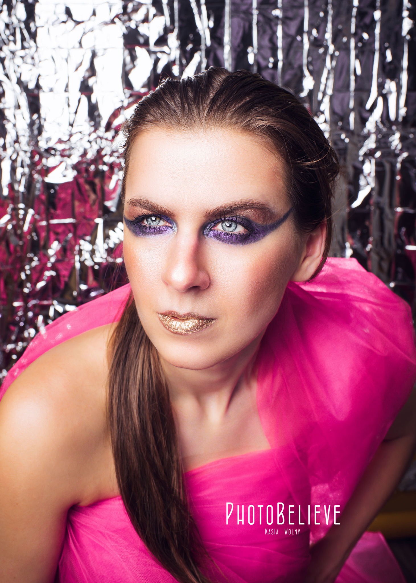 Portret kobiety z intensywnym makijażem oczu w odcieniach fioletu i złotymi ustami, ubrana w różową kreację na tle błyszczącej, srebrnej dekoracji.