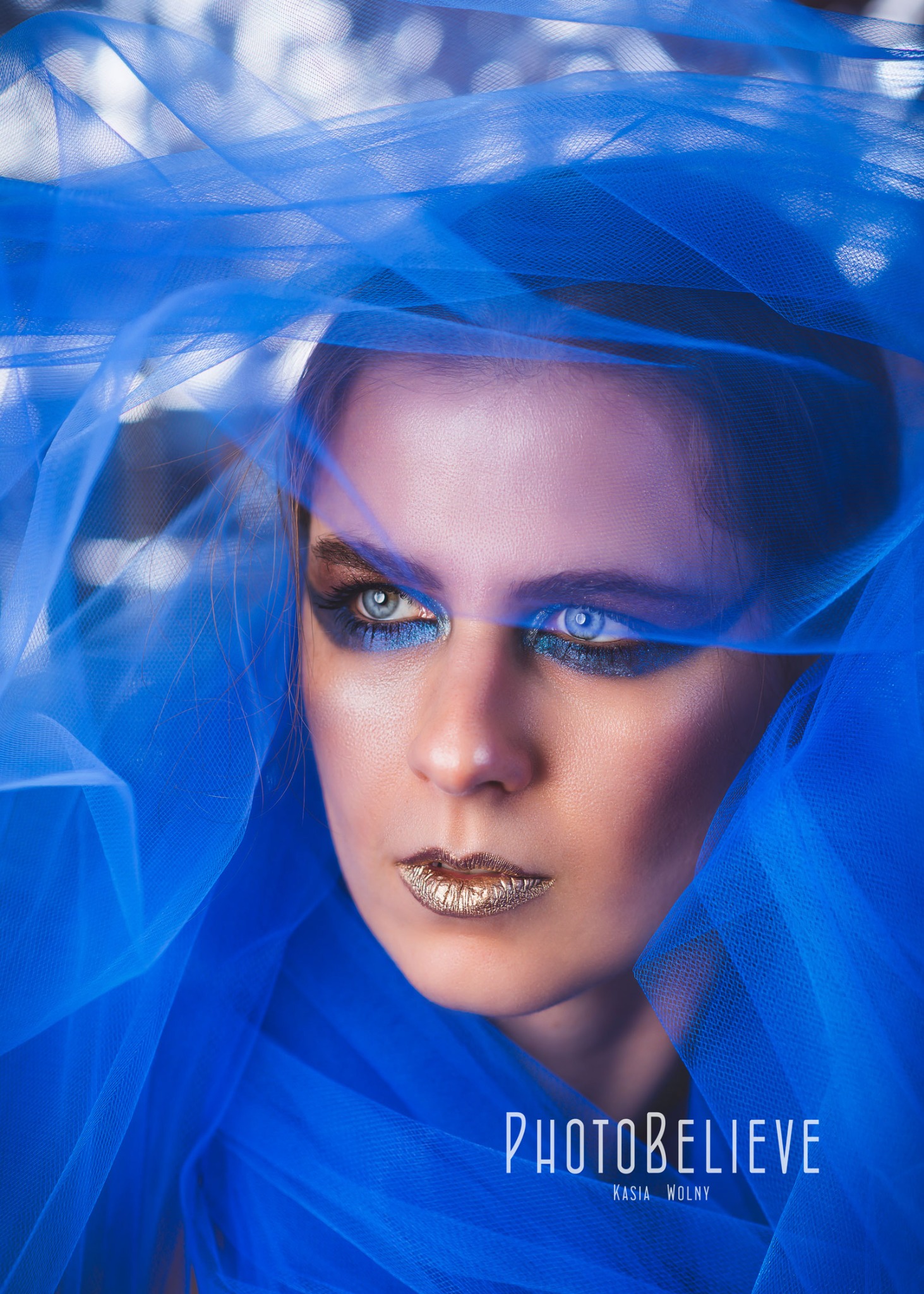 Portret kobiety z niebieską woalką, mocny makijaż oczu i złote usta, logo Photobelieve w dolnej części kadru.