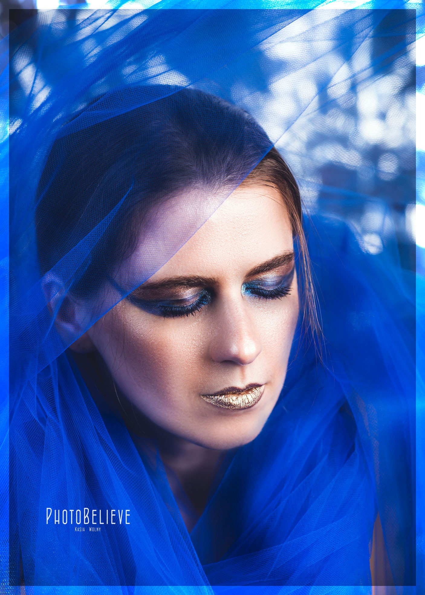 Portret kobiety z zamkniętymi oczami, okrytej niebieskim tiulem, z metalicznym makijażem oczu i złotymi ustami.