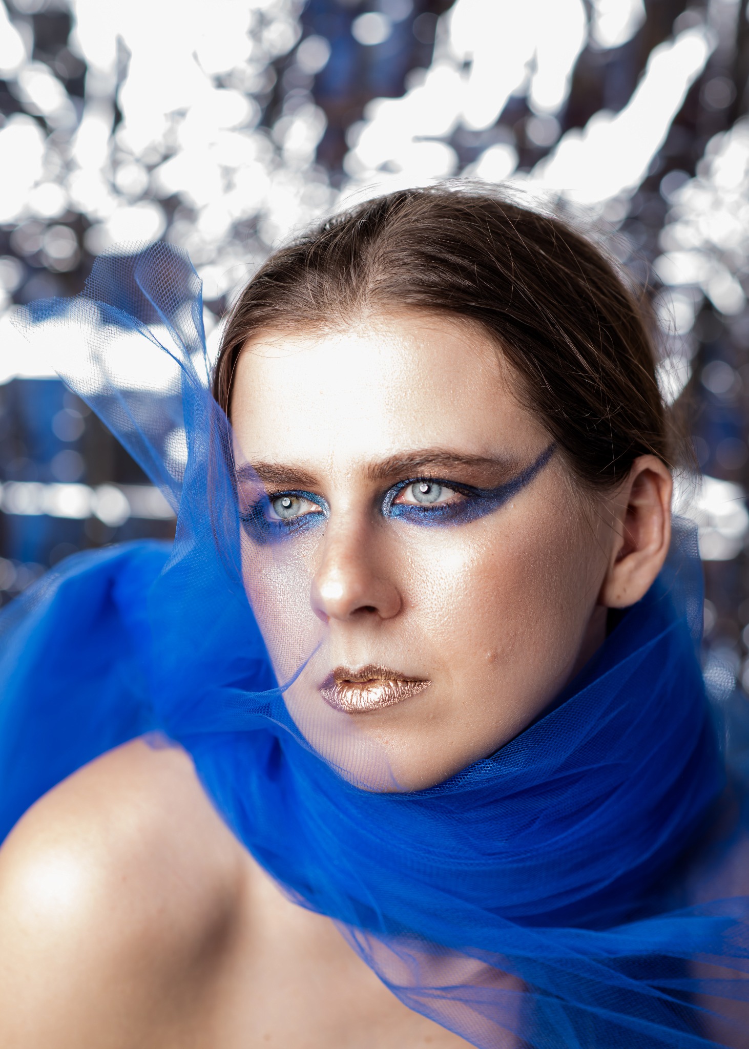 Portret kobiety z artystycznym makijażem w odcieniach niebieskiego i złotych ustach, owiniętej w błękitny tiul na tle srebrnego, rozmytego tła.