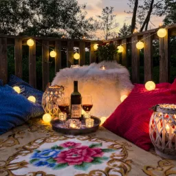 Romantyczny piknik na balkonie o zmierzchu: wino, świece, poduszki i girlanda świetlna, widok na zielone drzewa.