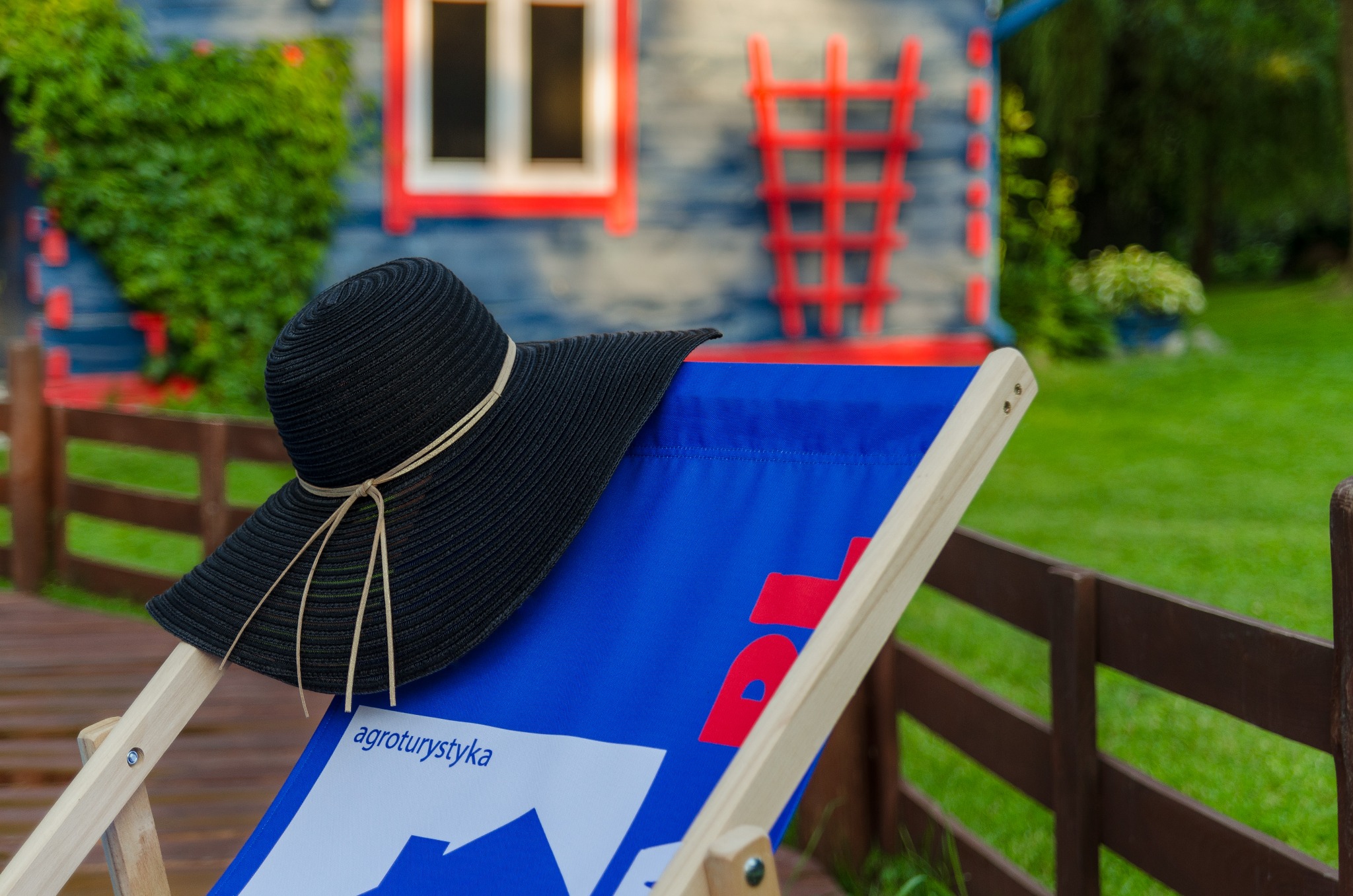 Czarny kapelusz na niebieskim leżaku z napisem 'agroturystyka' na tle kolorowego domku z czerwonymi elementami i zielenią.