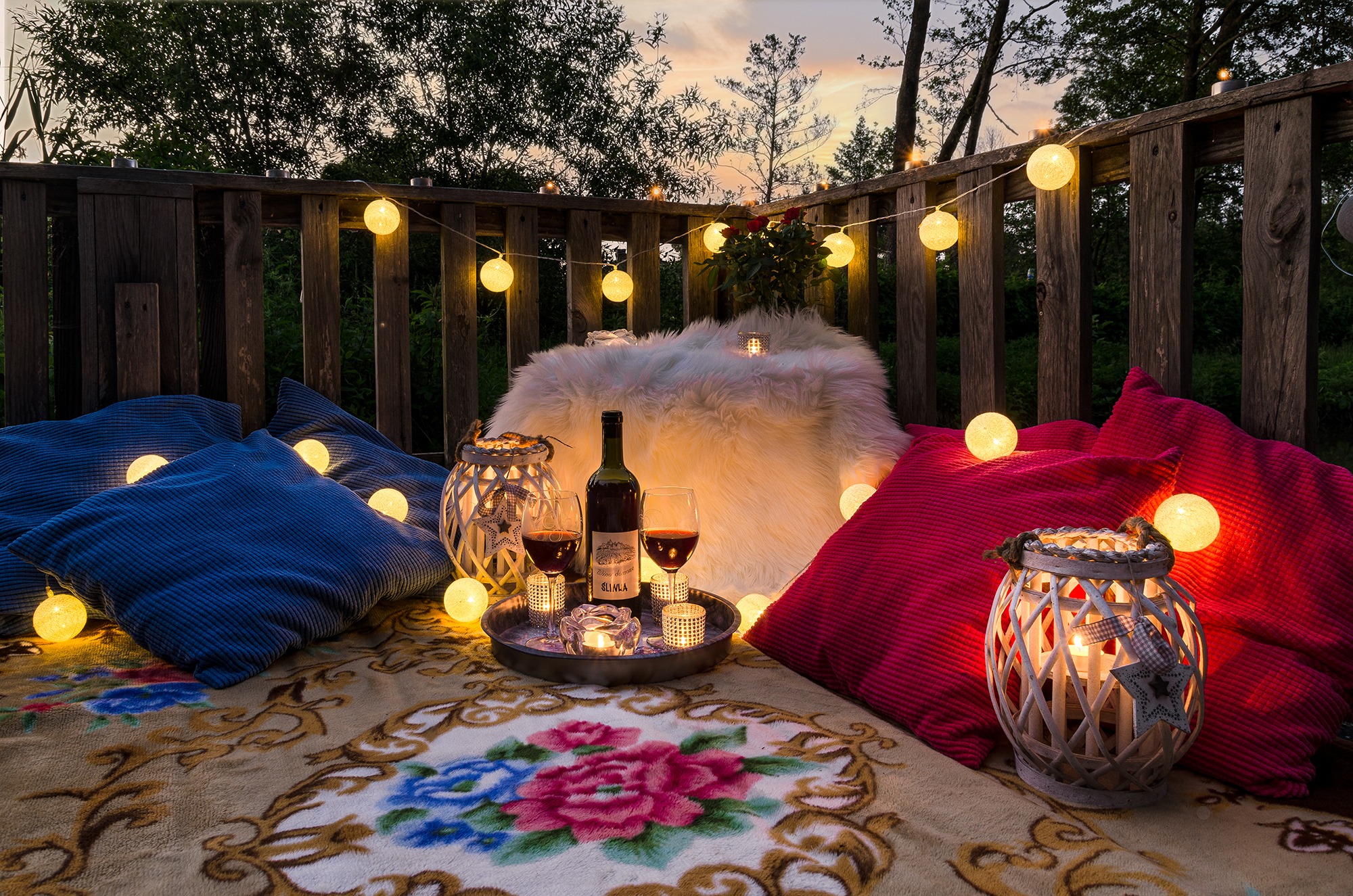 Romantyczny piknik na balkonie o zmierzchu: wino, świece, poduszki i girlanda świetlna, widok na zielone drzewa.