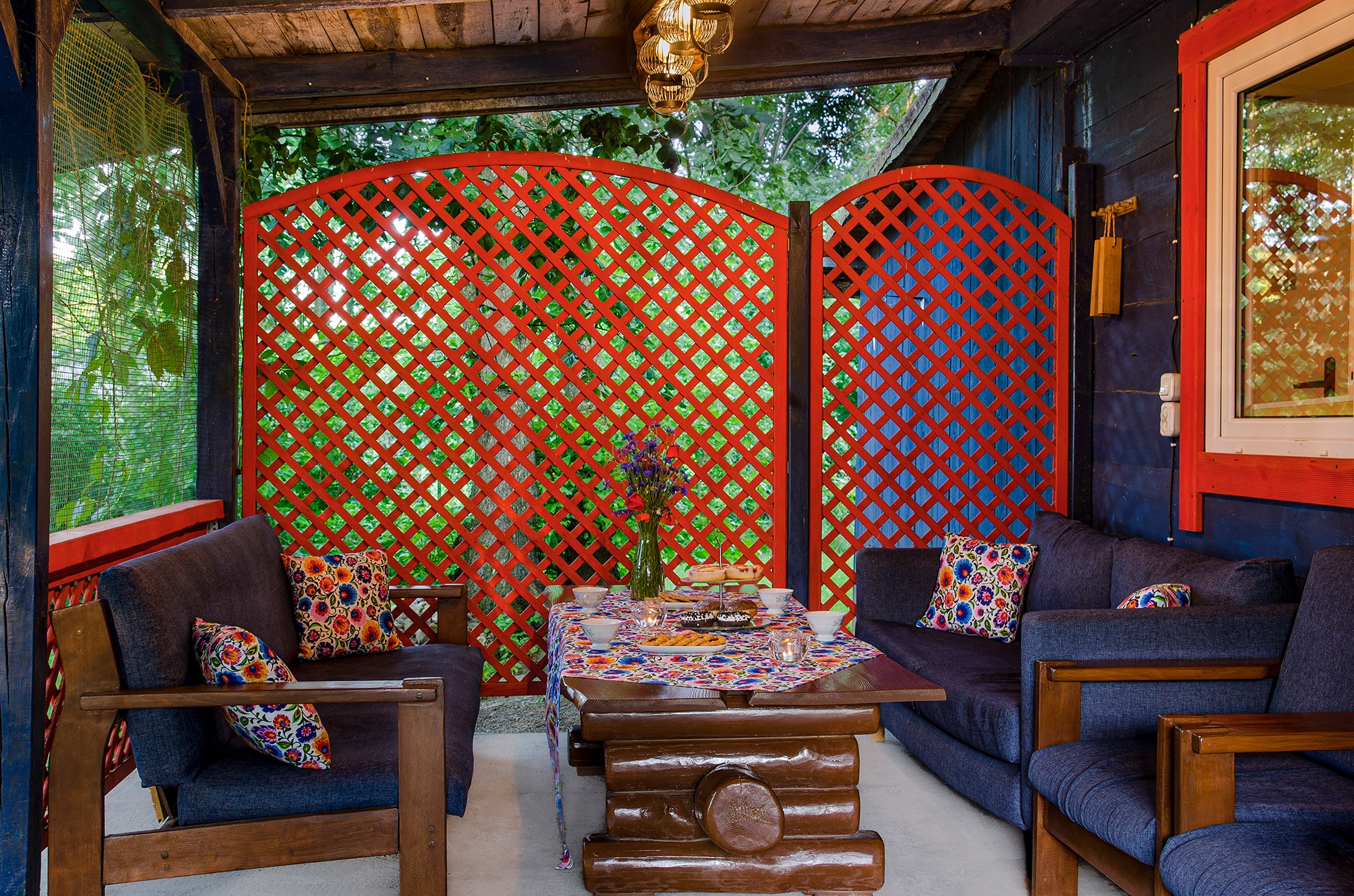 Zadaszony taras z drewnianymi meblami, sofami z kolorowymi poduszkami i stołem z przekąskami, czerwona ażurowa ściana w tle.