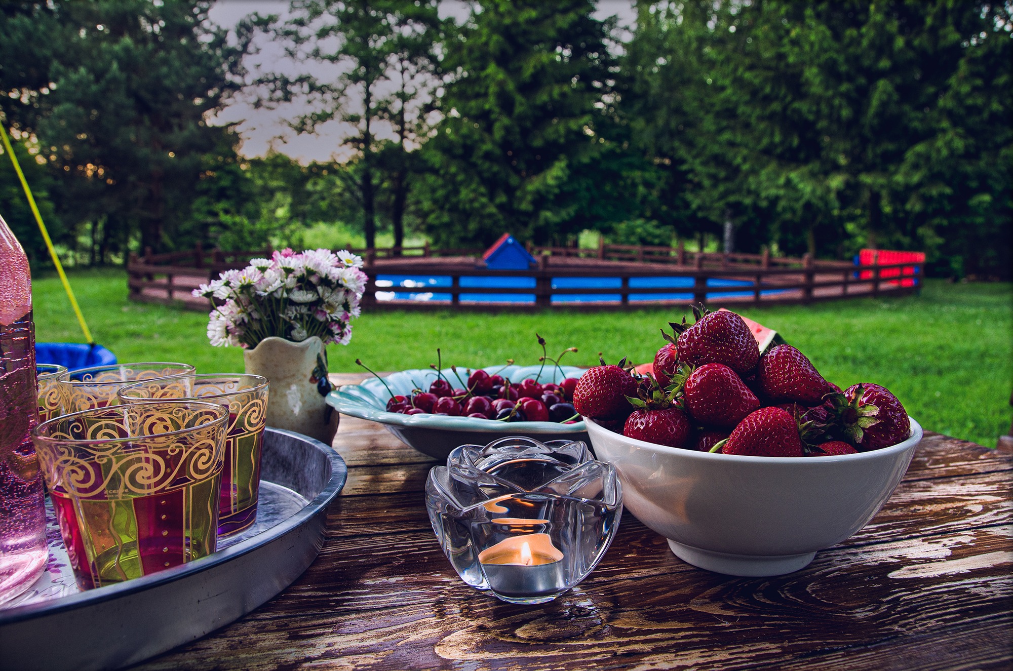 Letni stół z owocami: truskawki, wiśnie, napoje i kwiaty, w tle zielony trawnik i ogrodzenie basenu.