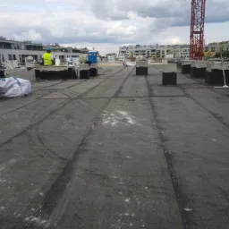 Prace dekarskie na płaskim dachu budynku wielorodzinnego, widoczny dekarz w żółtej kamizelce, materiały budowlane, elementy wentylacji i w tle żuraw budowlany na tle pochmurnego nieba.