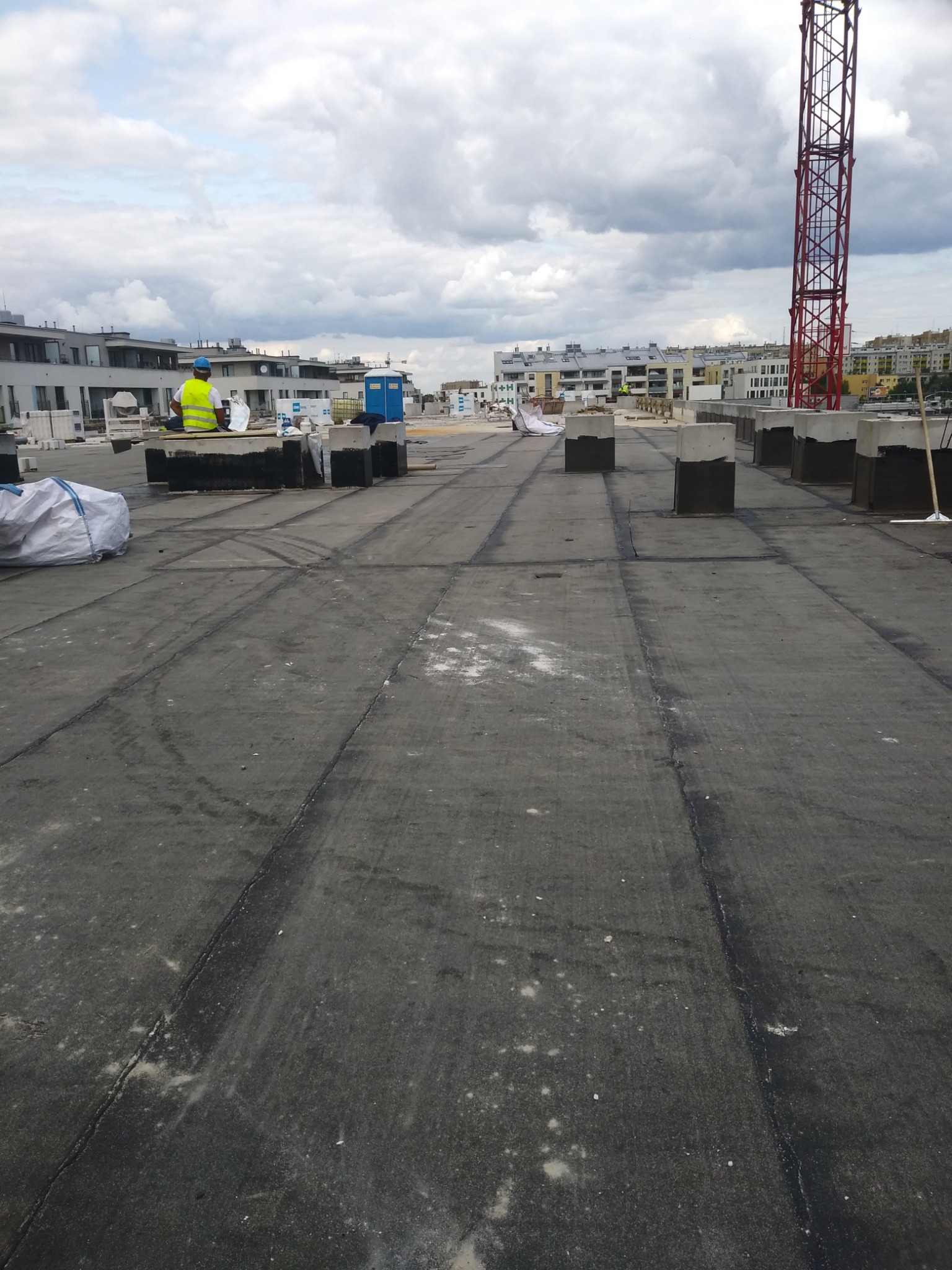 Prace dekarskie na płaskim dachu budynku wielorodzinnego, widoczny dekarz w żółtej kamizelce, materiały budowlane, elementy wentylacji i w tle żuraw budowlany na tle pochmurnego nieba.