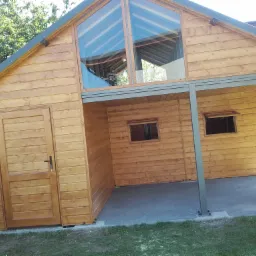 Drewniany budynek z dużym oknem trójkątnym, widoczna boczna ściana z drzwiami i zadaszona przestrzeń z dwoma oknami, całość na tle zieleni.