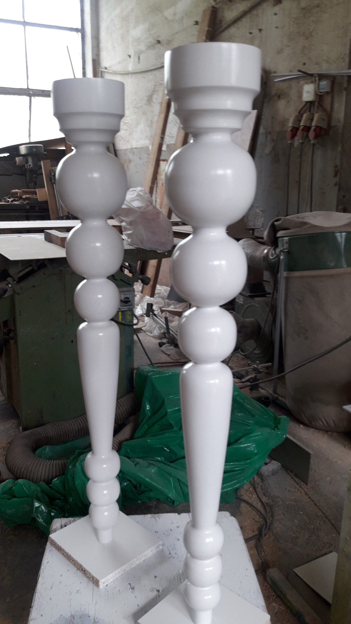 Dwie białe, toczone nogi meblowe z kulistymi elementami, stojące w warsztacie stolarskim, po świeżym malowaniu.