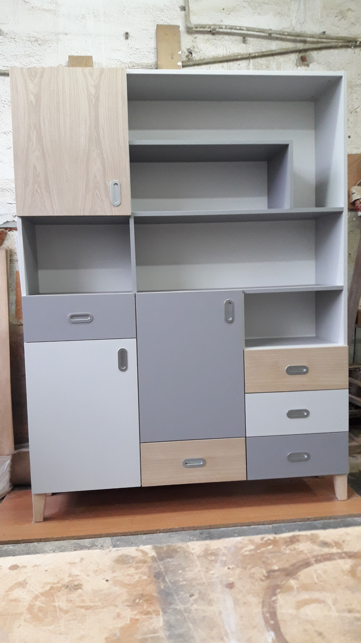 Wielobarwna, modułowa szafa z półkami i szufladami w odcieniach szarości i drewna, z podłużnymi uchwytami, ustawiona na podłodze w trakcie montażu.