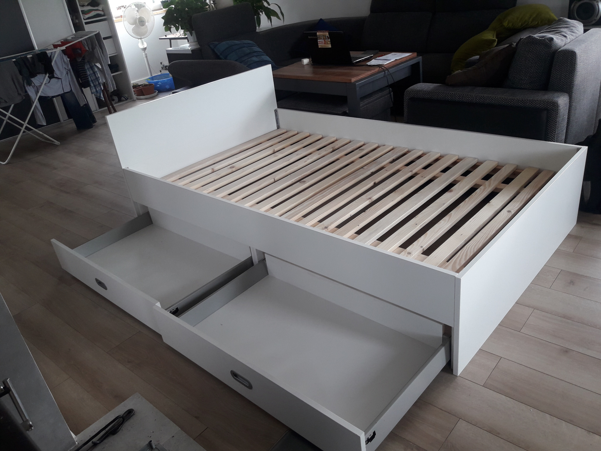 Białe łóżko z wysuwanymi szufladami na drewnianym stelażu, widoczne częściowo otwarte szuflady i fragment pokoju z suszarką na ubrania i sofą w tle.
