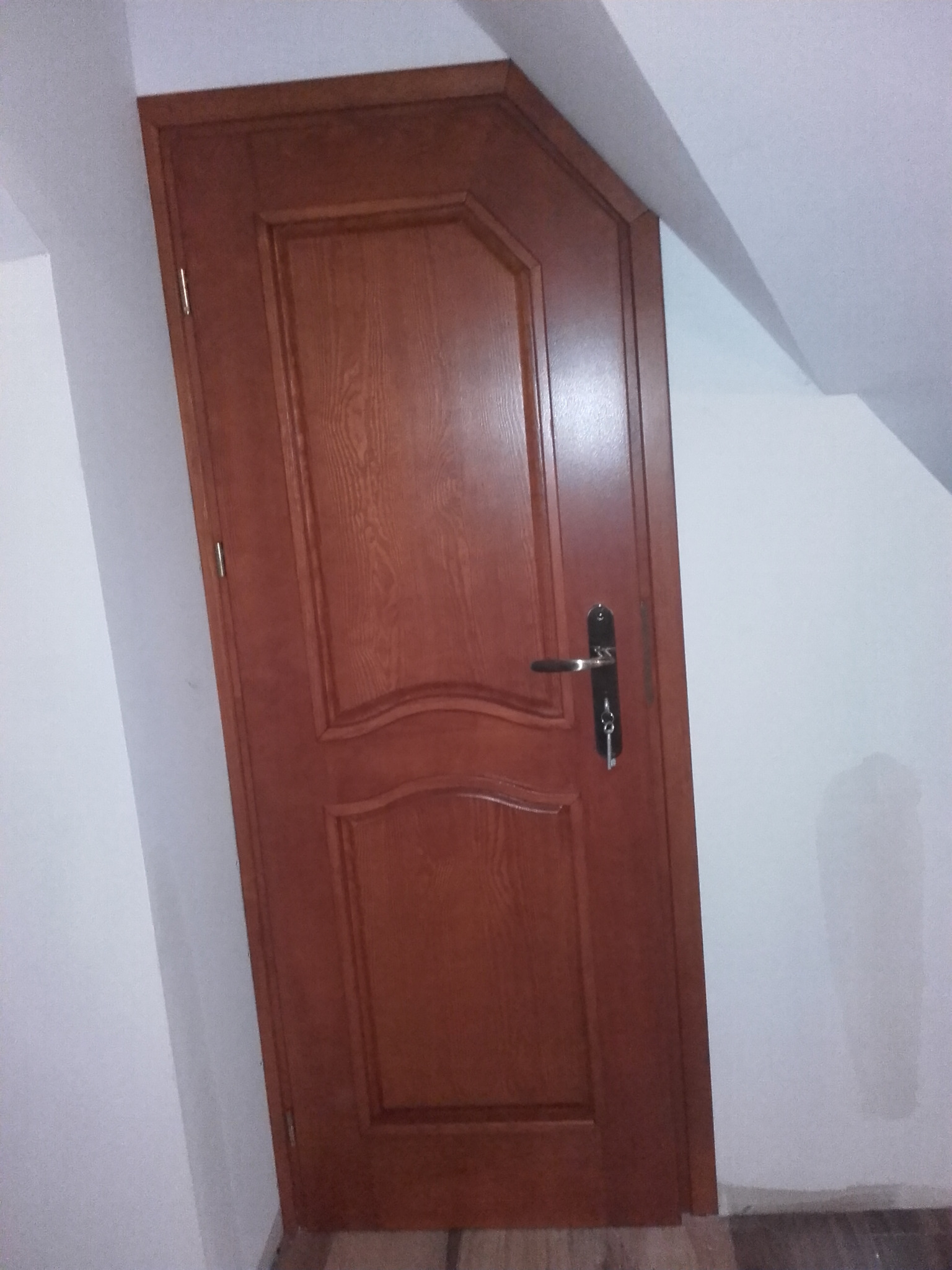Brązowe drzwi wewnętrzne z klamką i kluczem w ościeżnicy, zamontowane w pomieszczeniu ze skośnym sufitem.