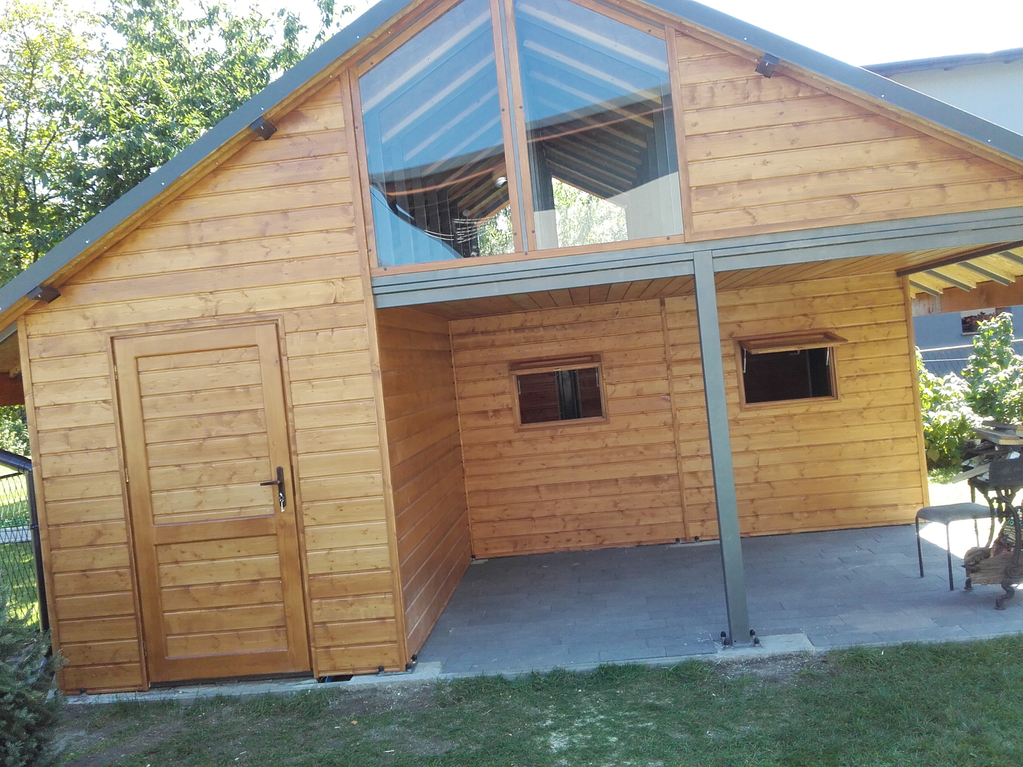 Drewniany budynek z dużym oknem trójkątnym, widoczna boczna ściana z drzwiami i zadaszona przestrzeń z dwoma oknami, całość na tle zieleni.