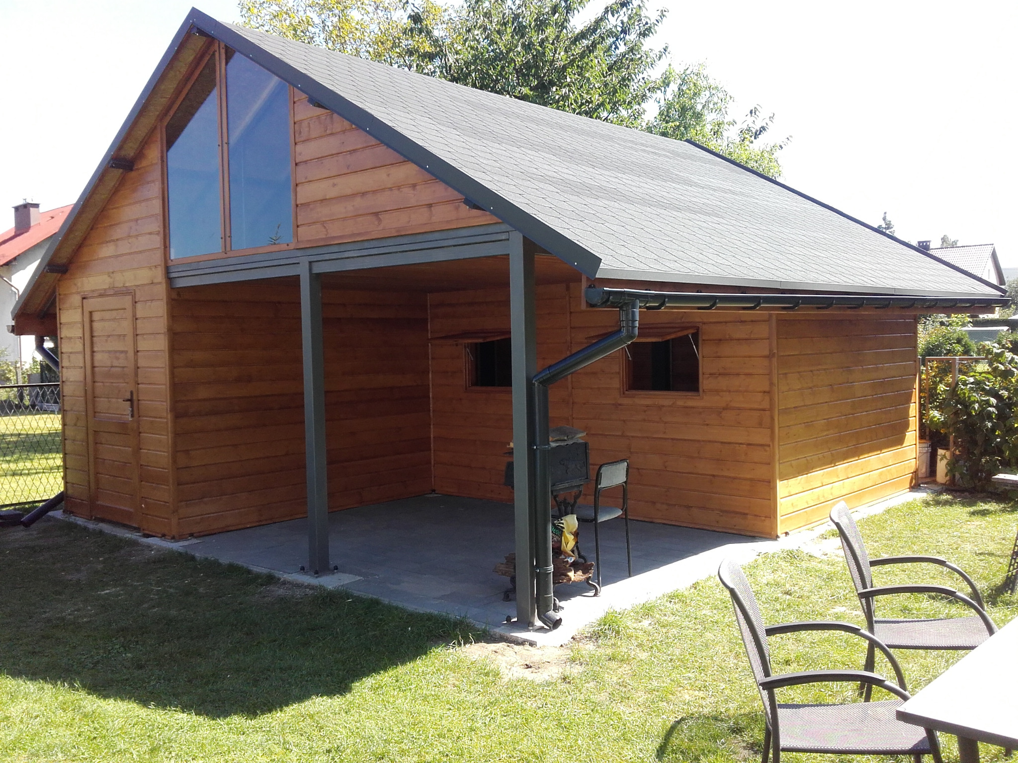 Drewniana altana z zadaszeniem i przeszkleniem trójkątnym, z miejscem do grillowania i meblami ogrodowymi na trawniku.