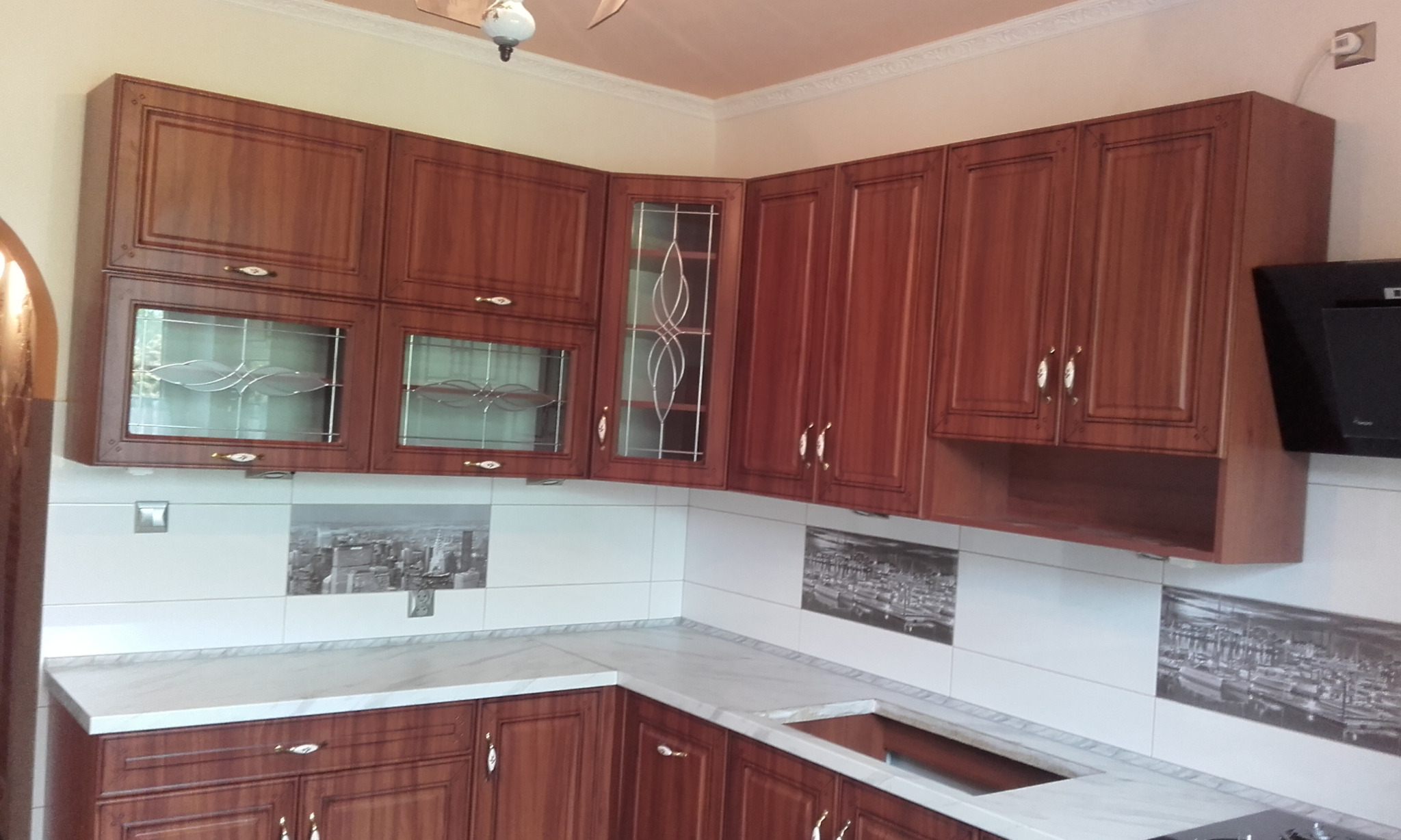 Brązowe szafki kuchenne z witrynami i dekoracyjnymi uchwytami, biały blat imitujący marmur, płytki z motywem miejskim na ścianie.