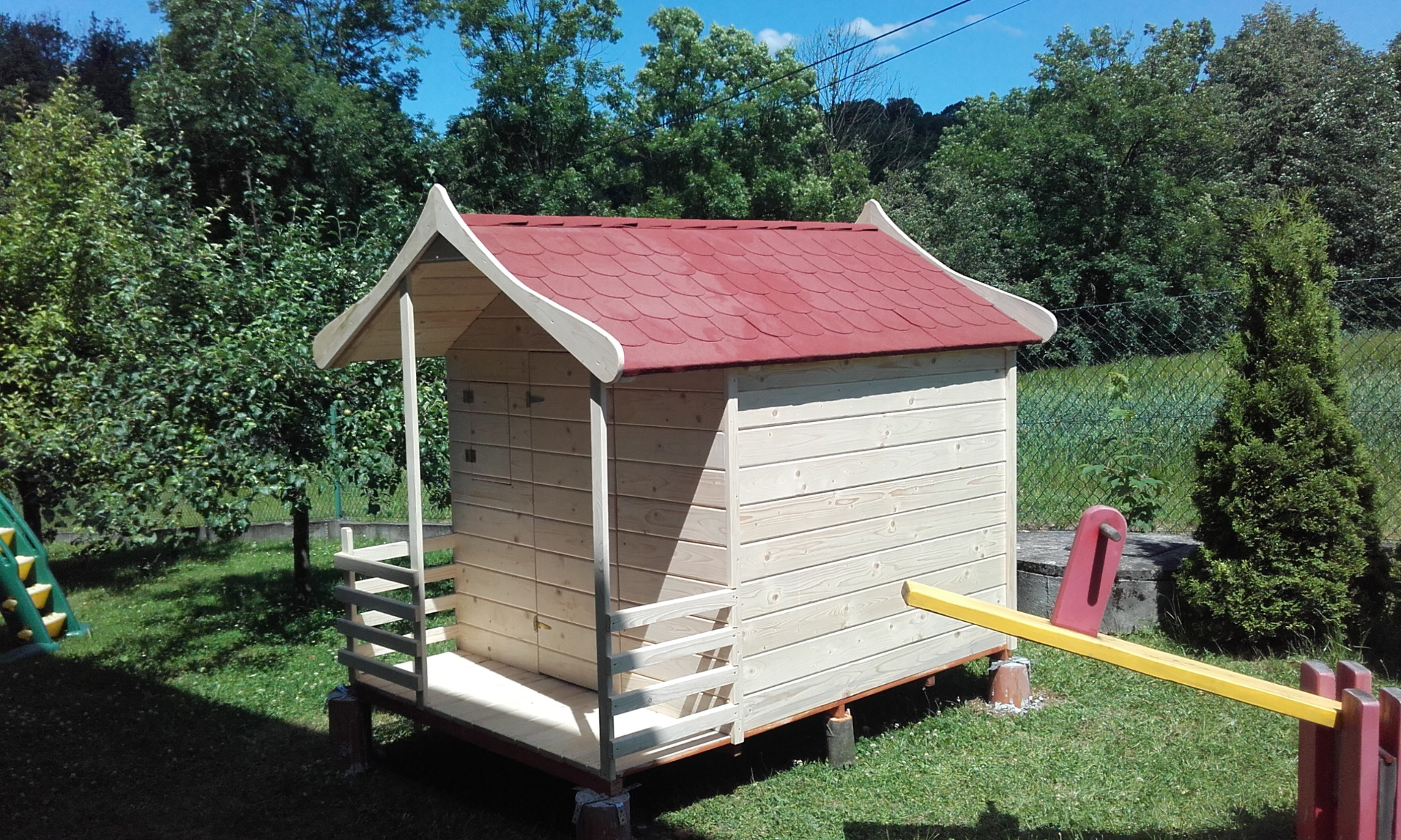 Mały drewniany domek dla dzieci z czerwonym dachem, stojący na trawie obok huśtawki, otoczony zielenią.