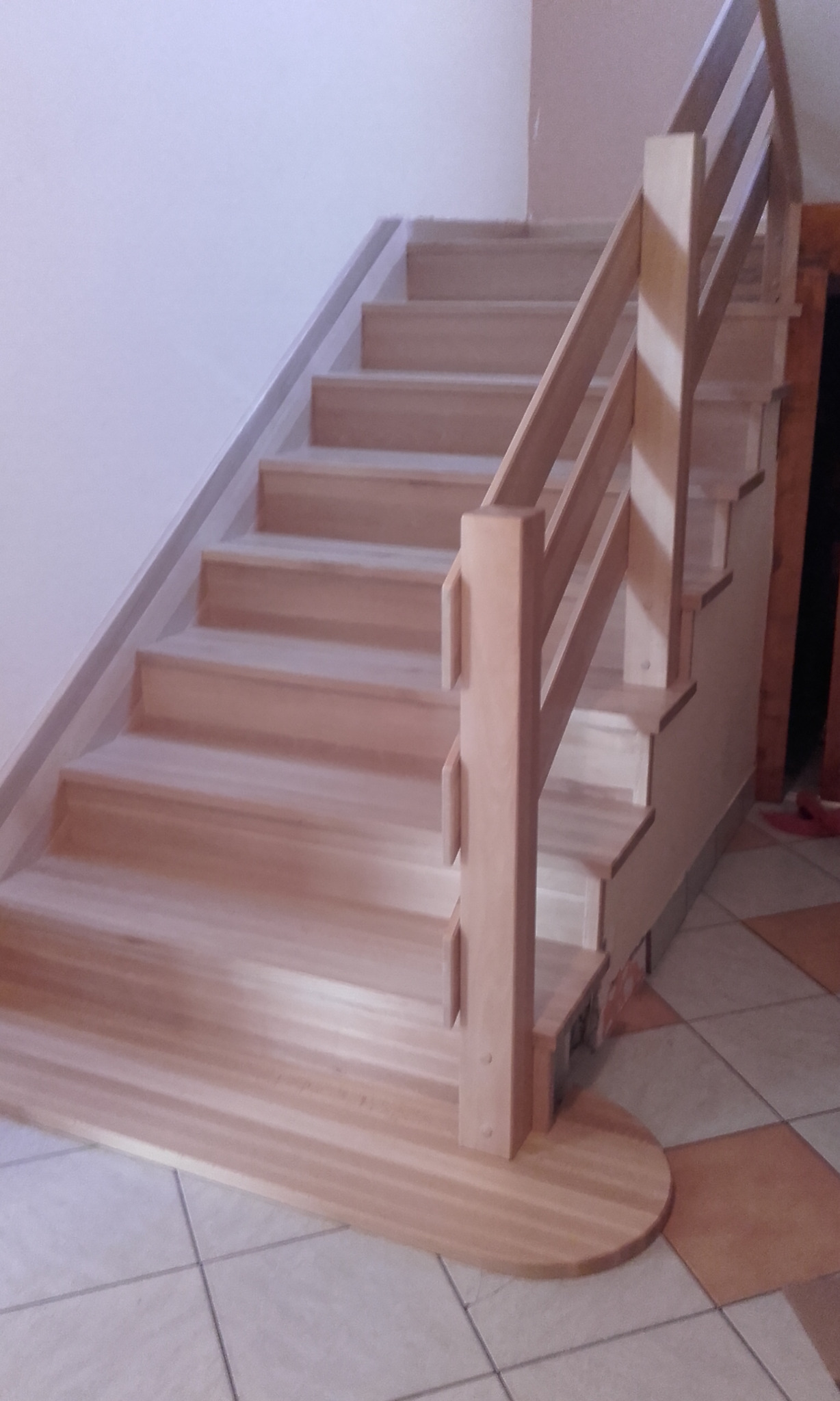 Jasne drewniane schody z prostą balustradą, widok z dołu na pierwsze stopnie z zaokrąglonym wykończeniem, na podłodze płytki ceramiczne.