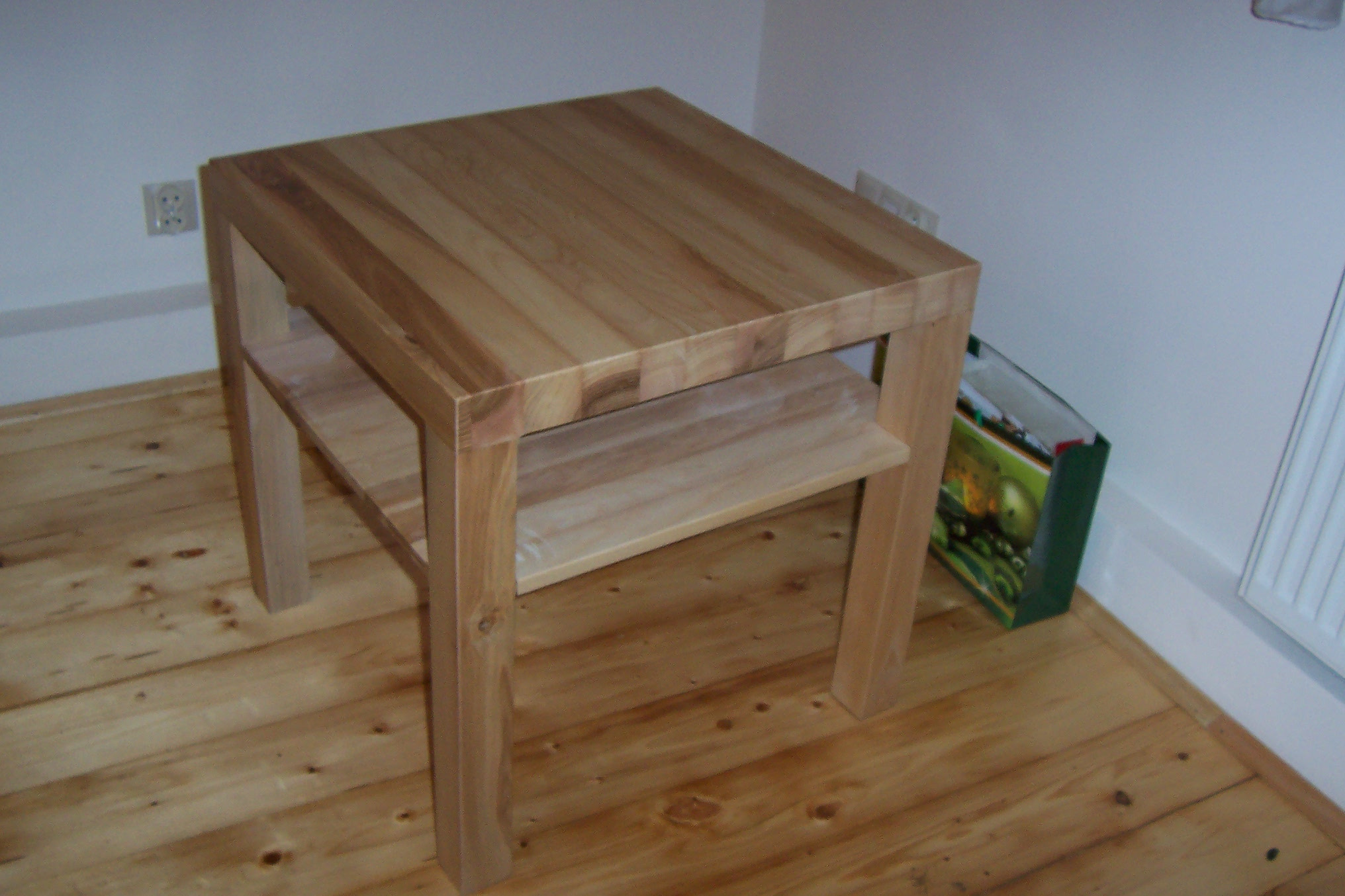 Surowy, nielakierowany stolik kawowy z jasnego drewna, z dodatkową półką, na tle drewnianej podłogi i białej ściany z gniazdkiem elektrycznym.