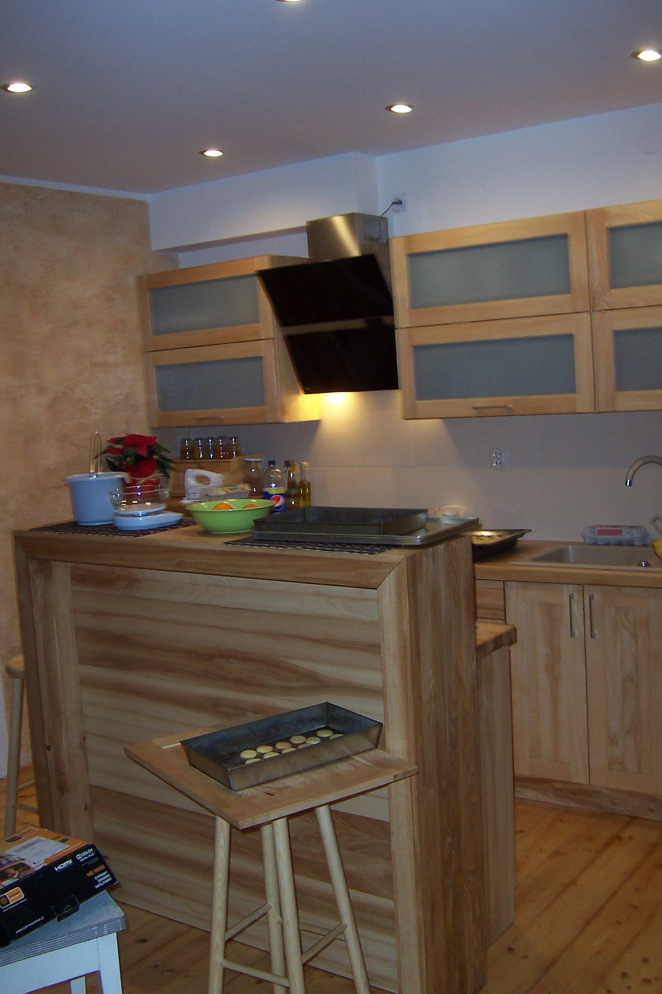 Jasna kuchnia z drewnianymi frontami szafek i barkiem, blacha z drobnymi ciasteczkami na stołku barowym, widoczny okap i oświetlenie punktowe.