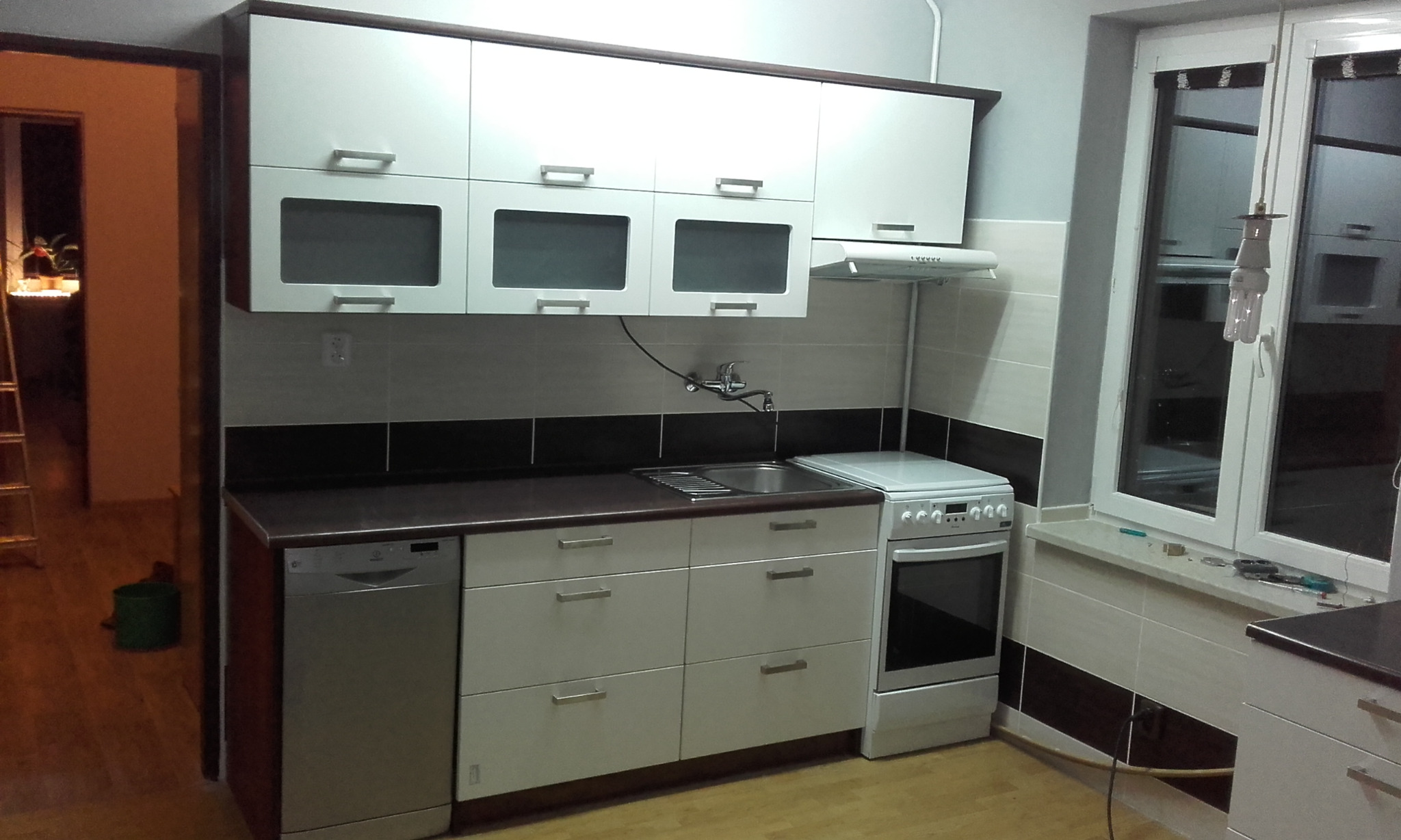 Nowoczesna, jasna kuchnia z białymi frontami szafek i ciemnym blatem, widok na zlewozmywak, kuchenkę i okno z akcesoriami montażowymi na parapecie.