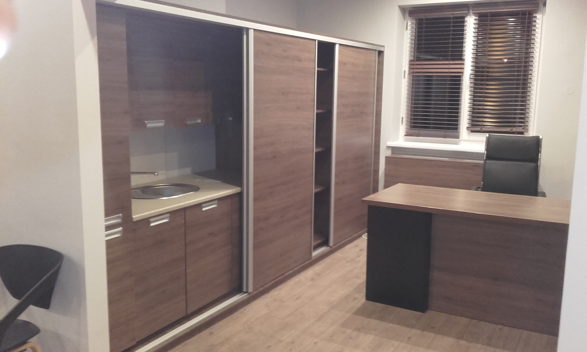 Wnętrze z zabudową kuchenną z umywalką, szafą przesuwną z półkami oraz biurkiem z krzesłem na tle okna z drewnianymi żaluzjami.