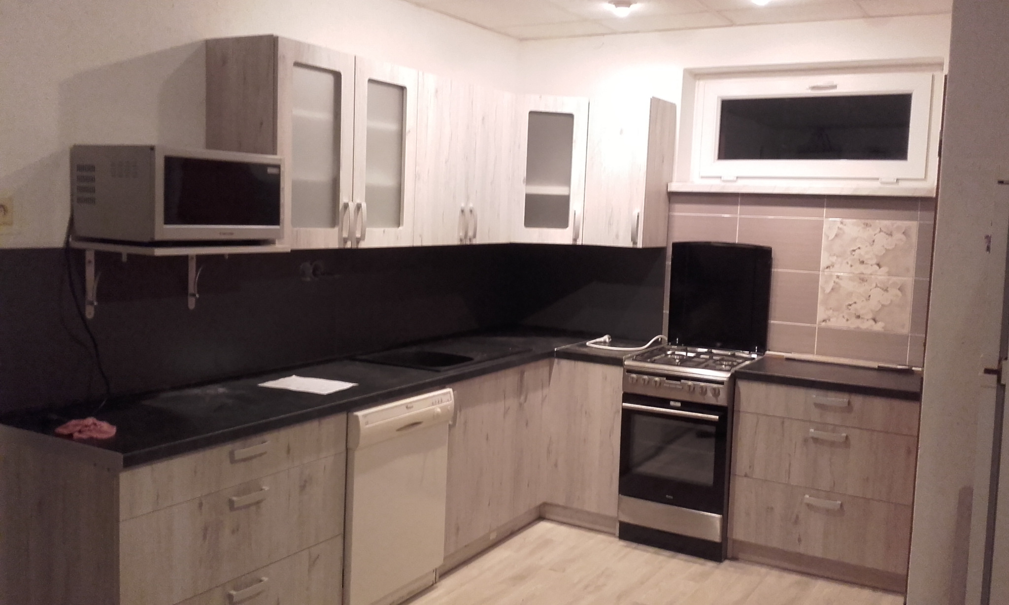 Jasna kuchnia z drewnopodobnymi frontami, czarnym blatem i zabudowaną kuchenką gazową; mikrofalówka na półce ściennej.