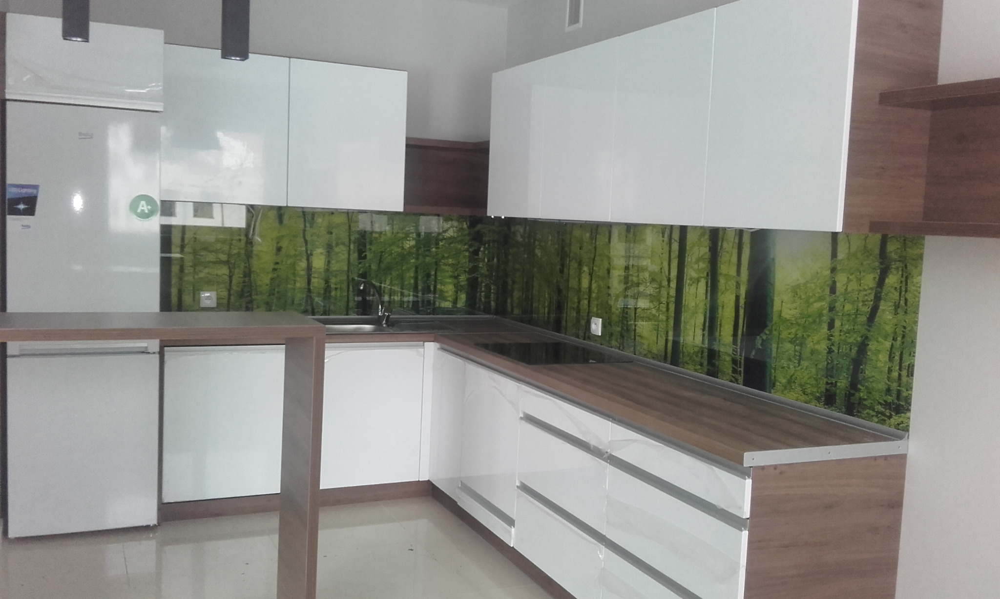 Nowoczesna kuchnia z białymi frontami i blatem w kolorze drewna, z zielonym panelem szklanym z motywem leśnym, oświetleniem punktowym i sprzętem AGD.