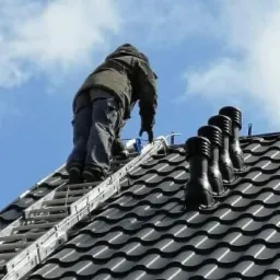 Pracownik na drabinie montuje elementy na dachu z czarnej dachówki falistej, widoczne cztery kominki wentylacyjne w rzędzie, błękitne niebo w tle.