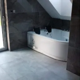 Nowoczesna łazienka na poddaszu z szarymi płytkami imitującymi kamień na ścianach i podłodze, biała narożna wanna z hydromasażem i oknem dachowym.