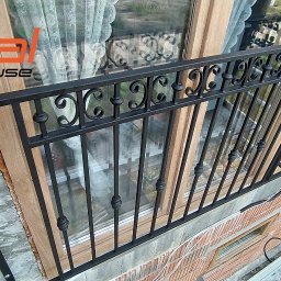 SMART METAL Tomasz Krause - Czarna, kuta balustrada okienna z dekoracyjnymi elementami w kształcie litery C, zamontowana na ceglanym murze budynku, widok z boku podczas montażu, drabina w tle.