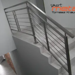 Szczegółowe ujęcie schodów z nowoczesną, metalową barierką z poziomymi prętami, oświetlonych z góry, z widocznym logo firmy 'smart metal Tomasz Krause' na ścianie.