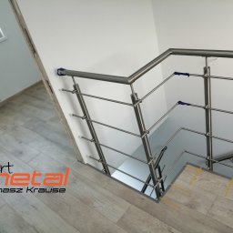 SMART METAL Tomasz Krause - Wnętrze domu z zamontowaną balustradą ze stali nierdzewnej przy schodach, widoczne zabezpieczenia na schodach i logo firmy w rogu zdjęcia.