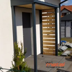 Zadaszenie nad wejściem do domu z drewnianymi lamelami po prawej stronie, wsparte na metalowych słupach w kolorze grafitowym, widoczne z zewnątrz budynku, z przodu mała roślina, w tle dom sąsiada.