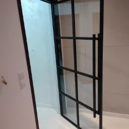 Otwarta, przeszklona konstrukcja z czarnymi profilami, pełniąca funkcję drzwi wewnętrznych, z widocznym fragmentem instalacji elektrycznej obok włącznika.