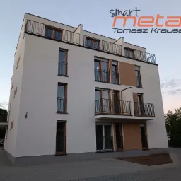 Nowoczesny budynek wielorodzinny z balkonami z czarnymi balustradami i balustradą na dachu, widziany z dołu, z logo firmy Smart Metal Tomasz Krause w prawym górnym rogu.