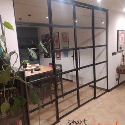 SMART METAL Tomasz Krause - Szklana ściana działowa z czarnymi metalowymi szprosami oddzielająca jadalnię, widoczny stół z nakryciem, w tle oświetlenie szynowe